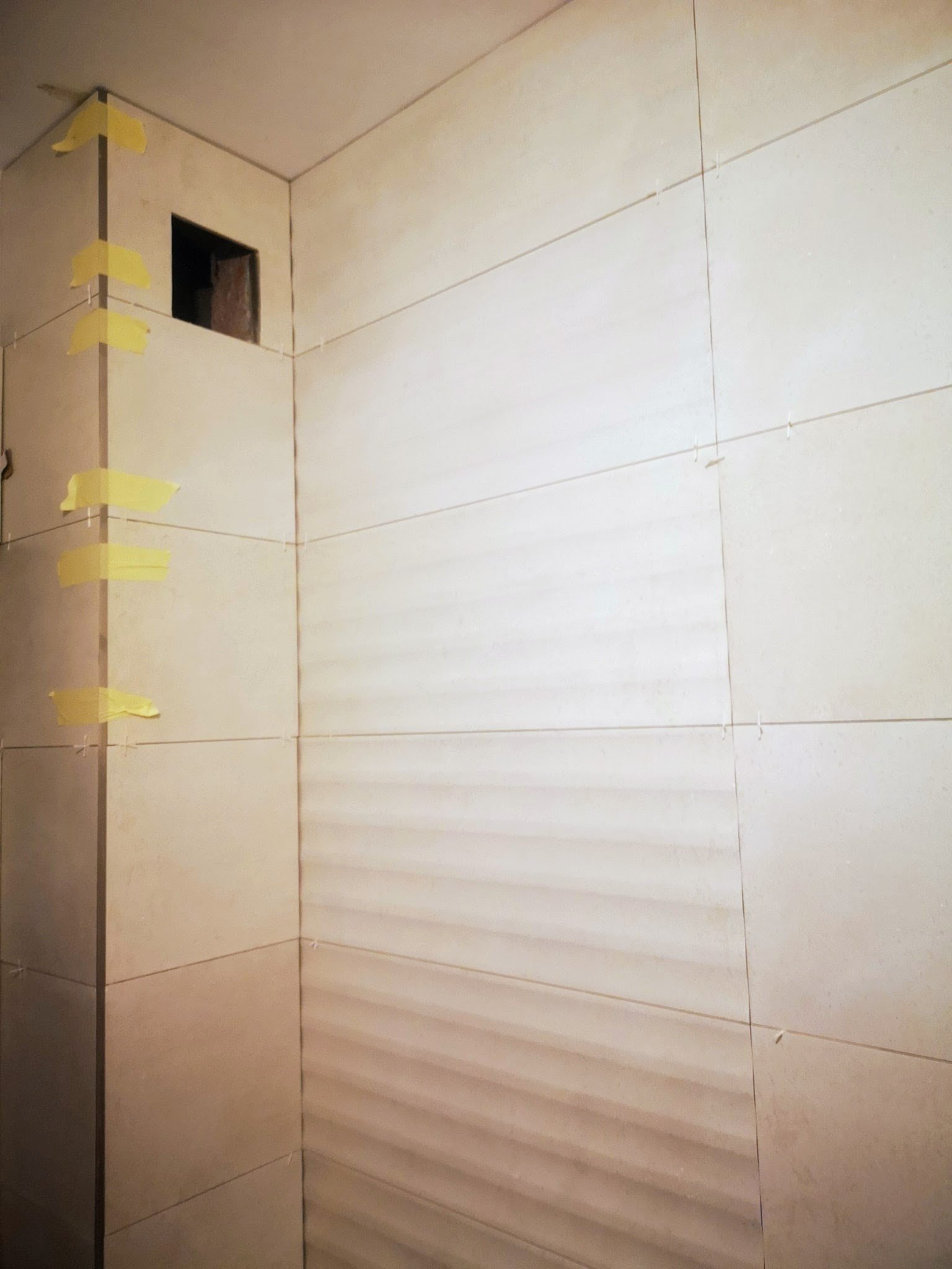 Interior of a building under construction with large beige tiles on the wall, a small square opening, and yellow tape marking the edges of tiles.