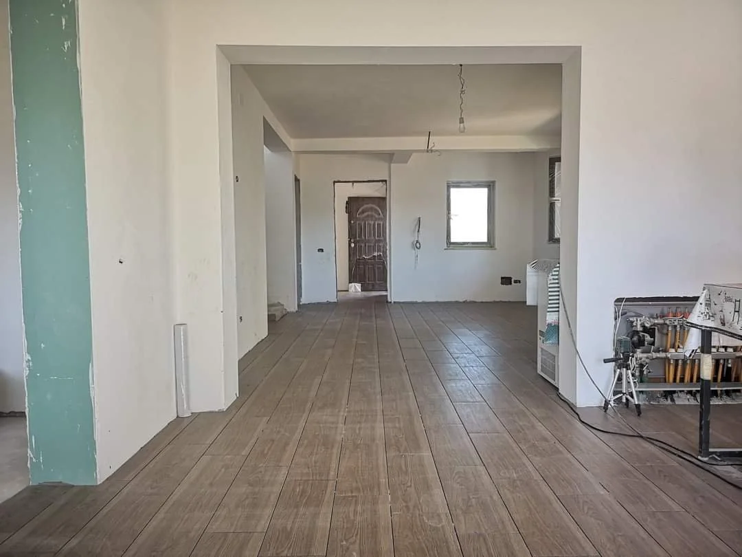 Empty room with new wooden flooring, white walls, and a visible doorway leading to the front door. Construction materials and tools are present, indicating ongoing renovation.