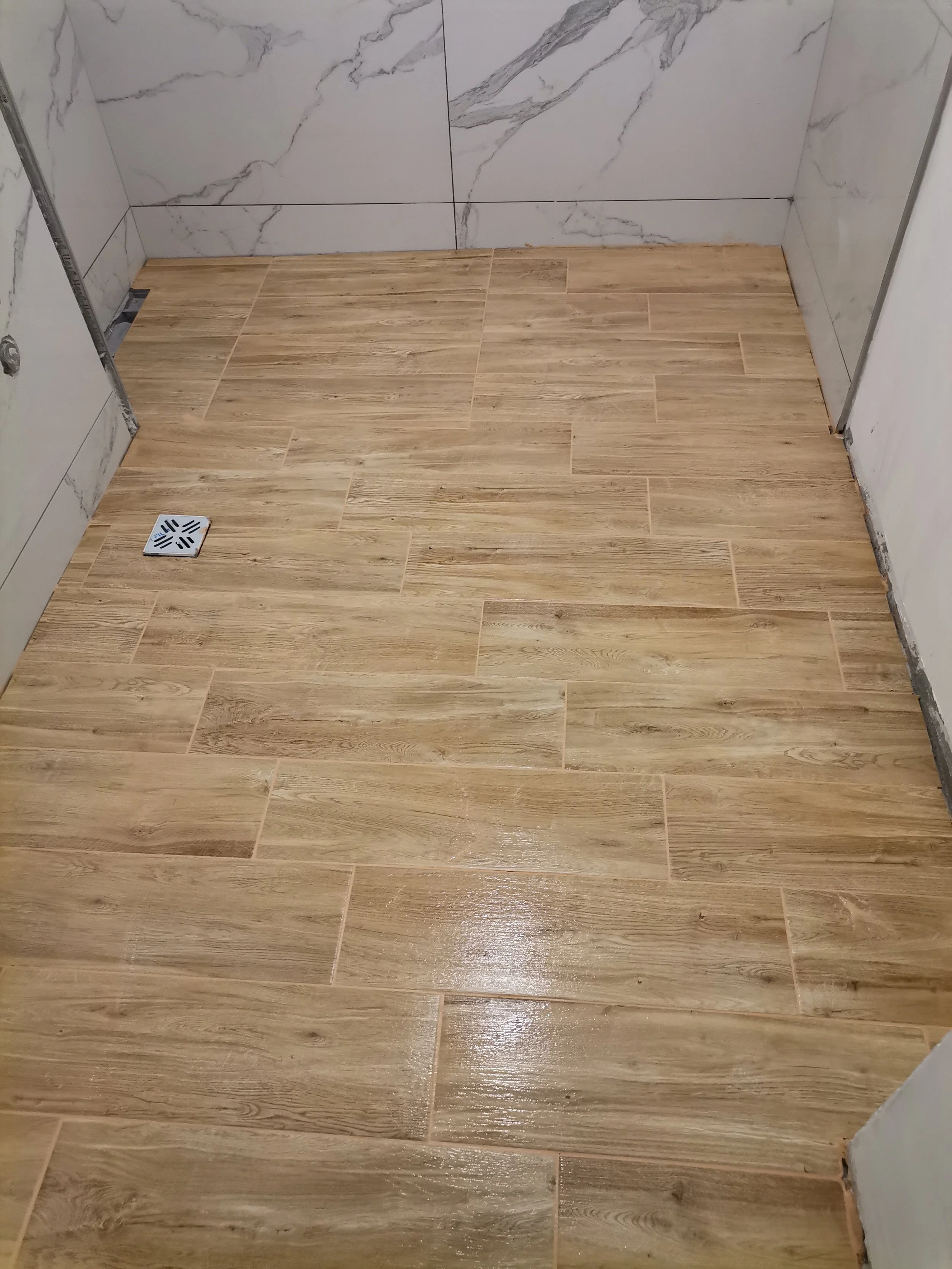 The image shows a newly installed wooden tile floor in a room, with a drain in the lower left corner and partially finished wall tiles on the adjacent walls.