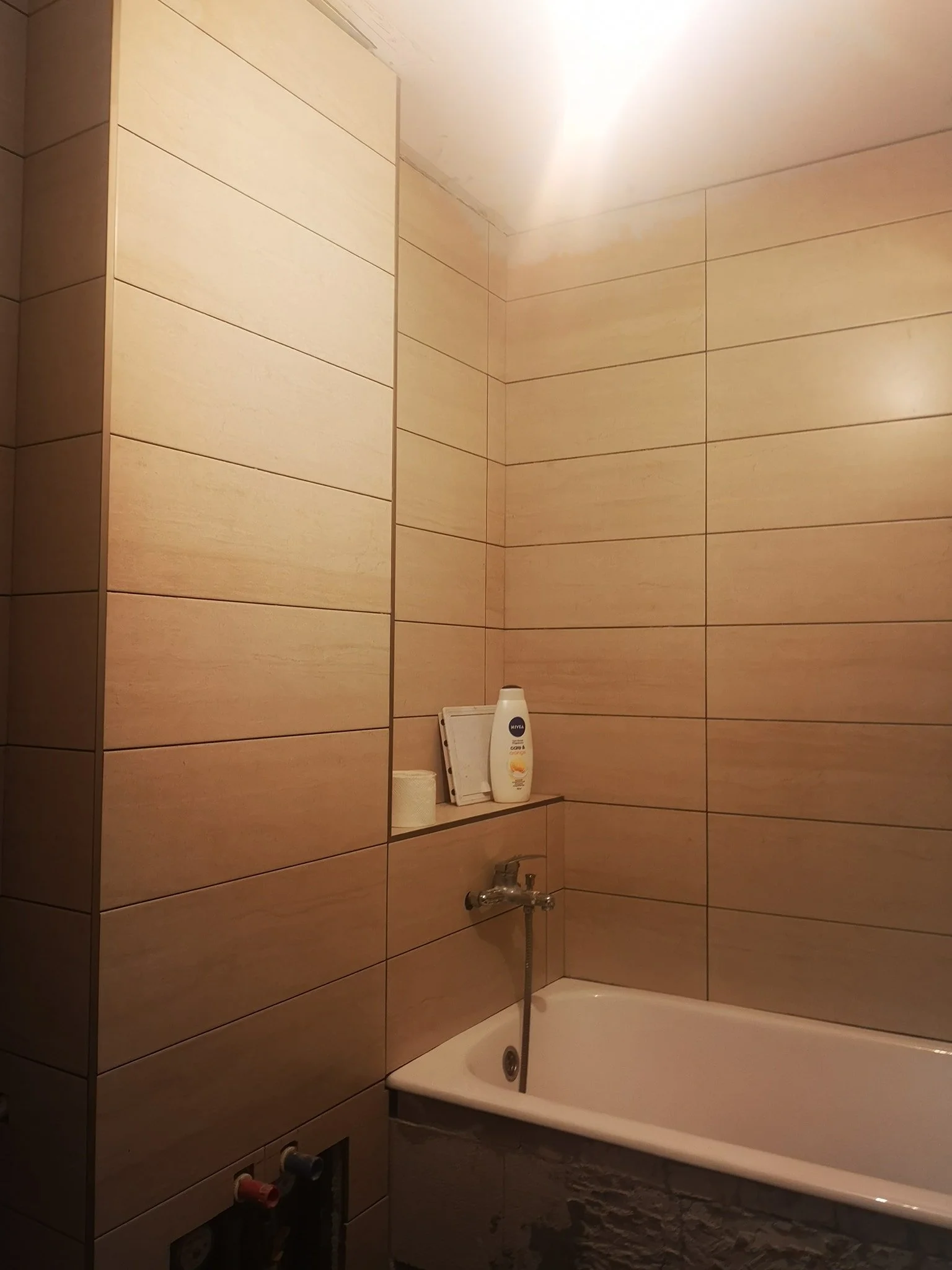 A bathroom with beige tiled walls. A white bathtub is visible with a chrome faucet and a handheld showerhead. On a built-in shelf above the bathtub, there are a bottle of lotion, some toilet paper, and a white frame. The bathroom appears to be modern