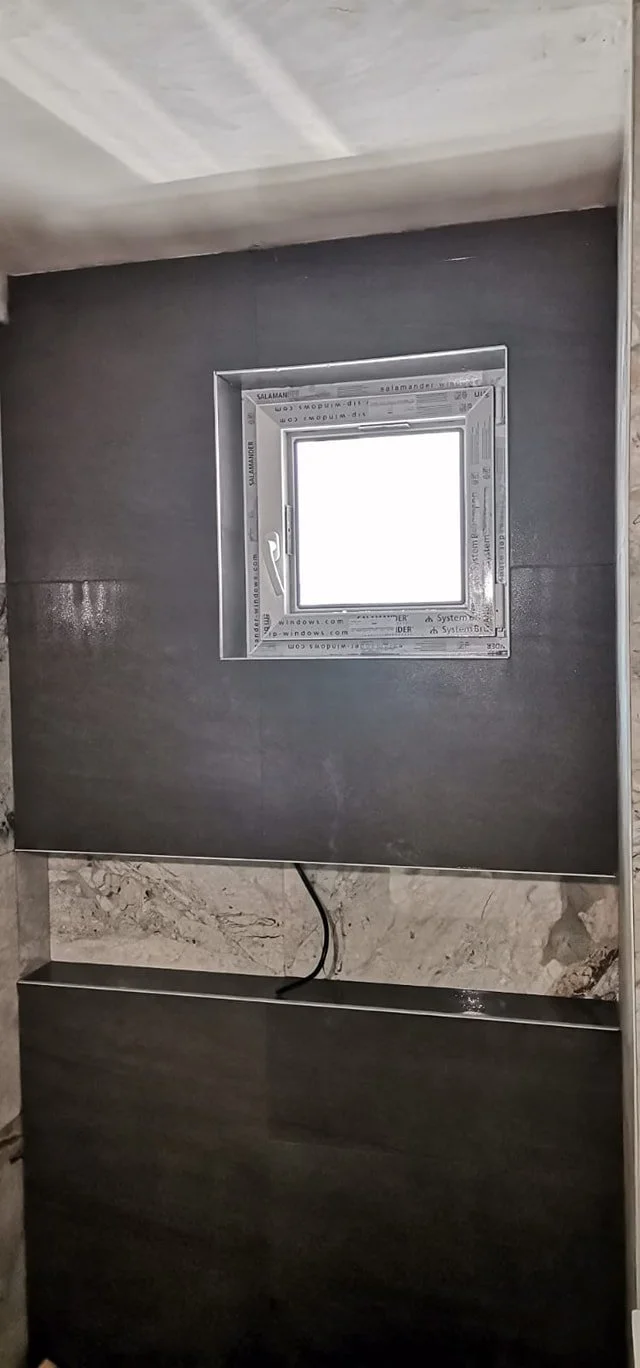 A small square window installed in a wall with dark gray tiling, surrounded by metallic framing.