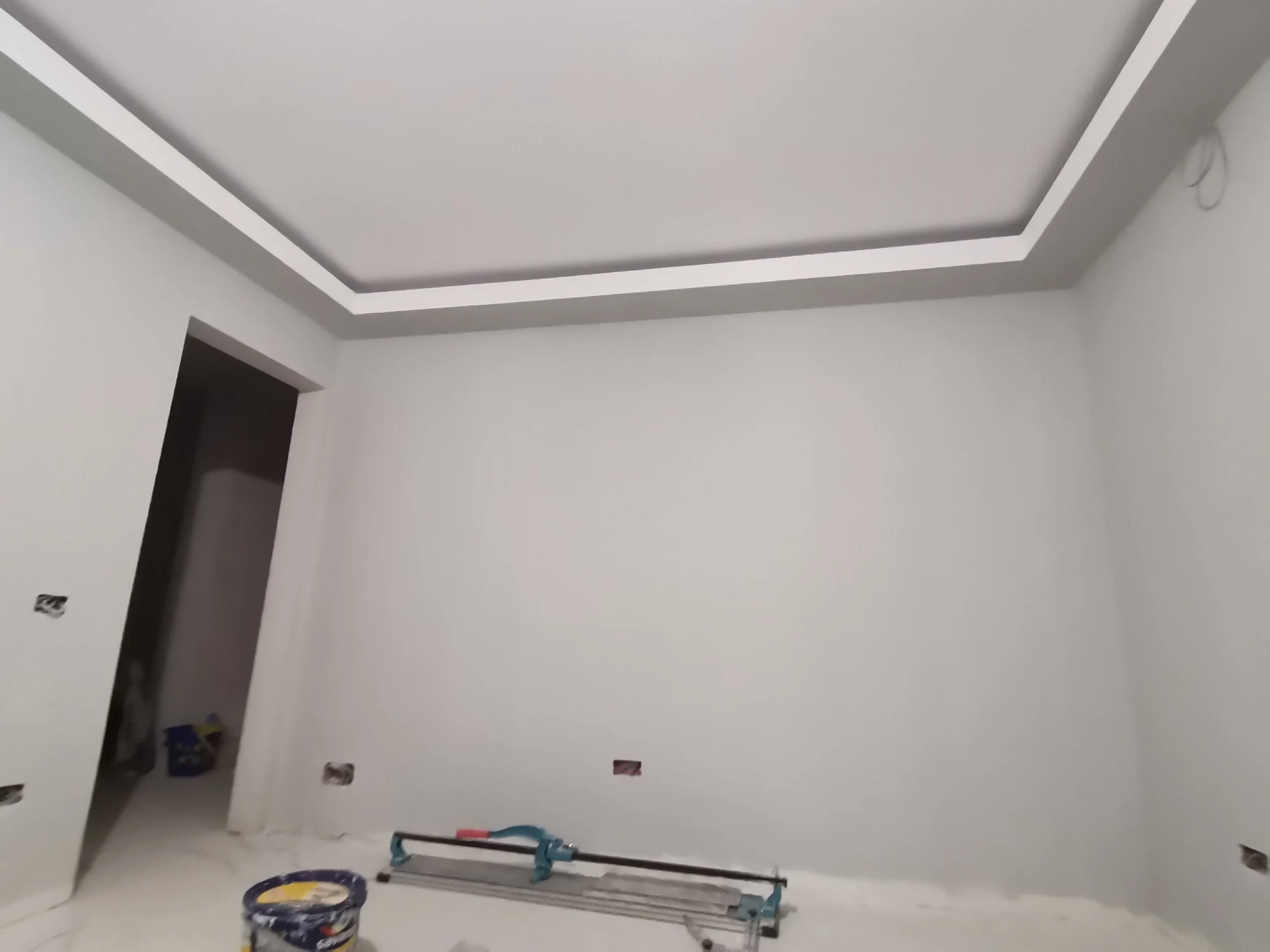 Empty white room with a tray ceiling, some construction equipment and supplies, including a stepladder, a paint bucket, and a scaffold in the corner.