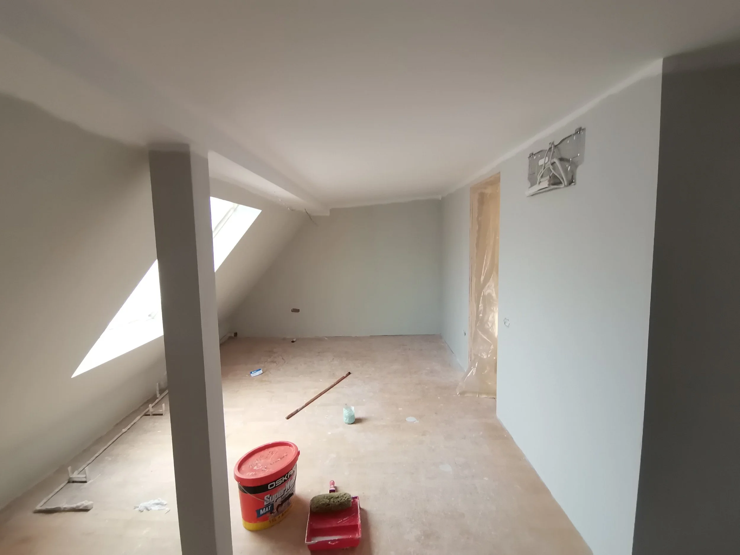 An unfinished room under construction with drywall, a slanted ceiling, and construction tools and materials on the floor, including a red bucket, a paint roller, and a small container, with wall openings and a window.