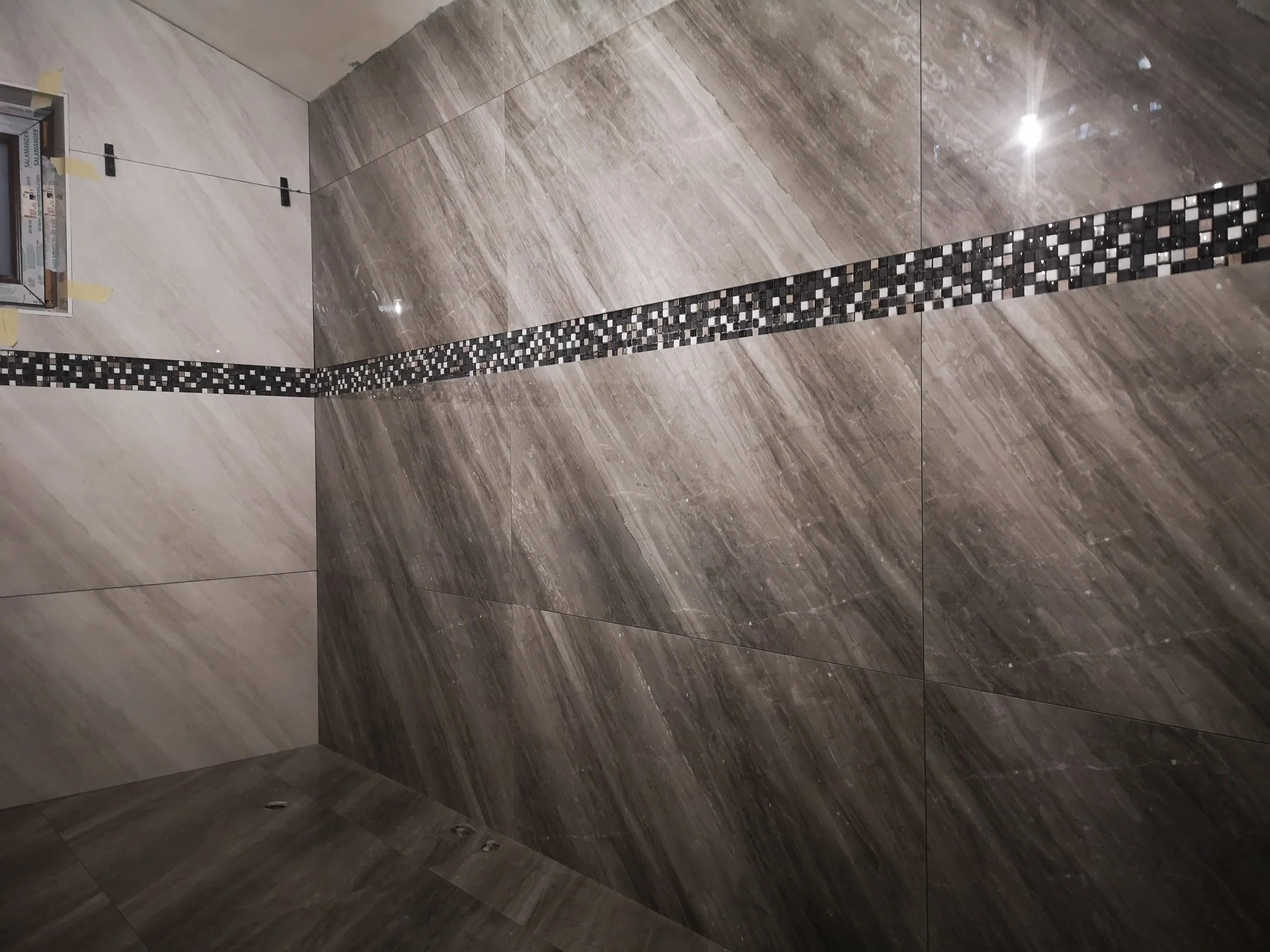 Bathroom with large brown and beige tiles on the wall and floor, a mosaic strip of small black, grey, and white tiles, and a vent near the bottom.