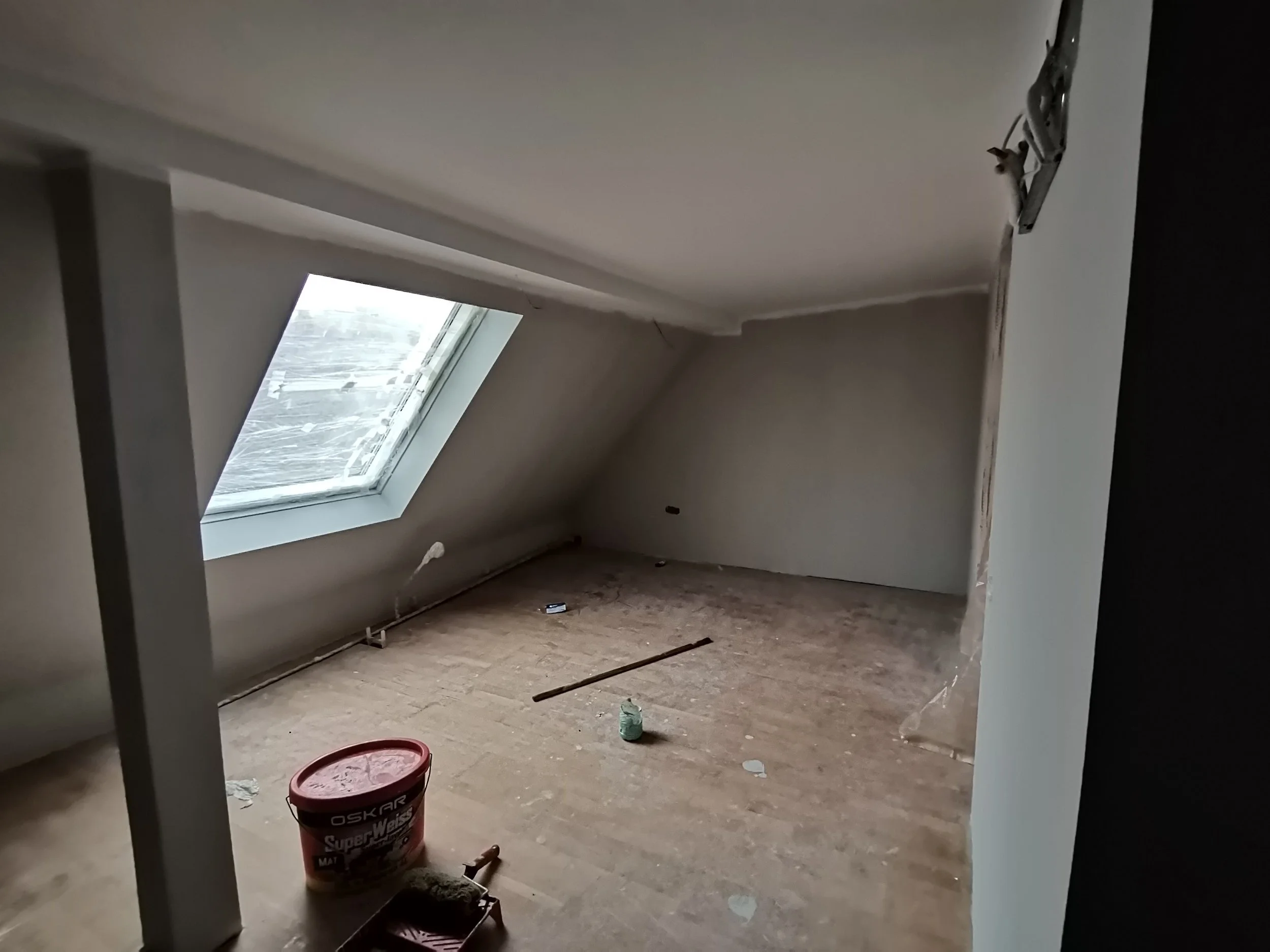 An unfinished attic room with a sloped ceiling and a large skylight window on the left. The floor is wooden and scattered with construction materials and tools, including a red bucket, a roll of tape, and some debris.