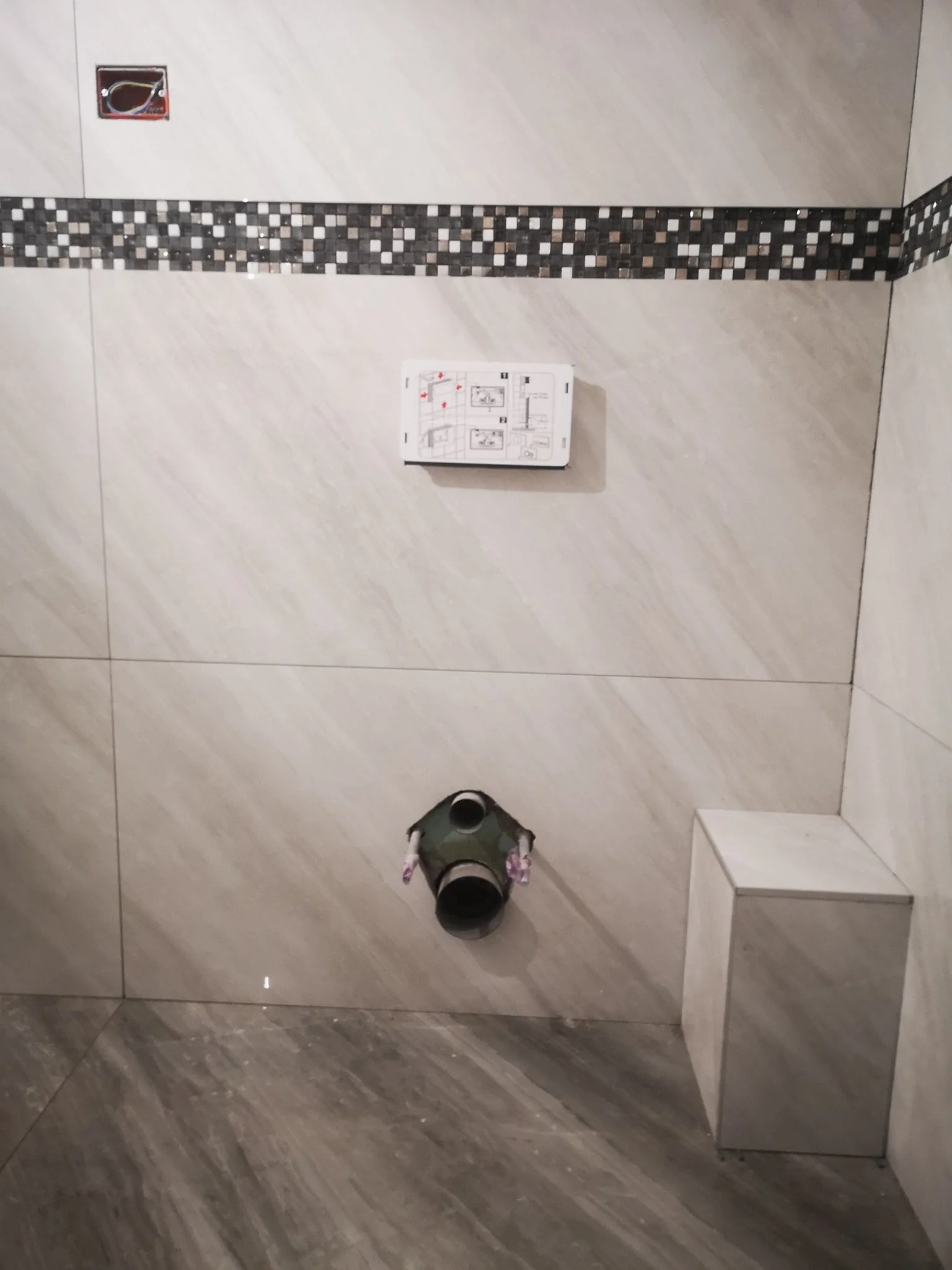 A mirror reflects a bathroom wall with beige tiles and a decorative strip of small, square, black, gray, and white mosaic tiles. There is an electrical outlet without a cover in the upper left corner. A small, white, rectangular instruction sheet or 