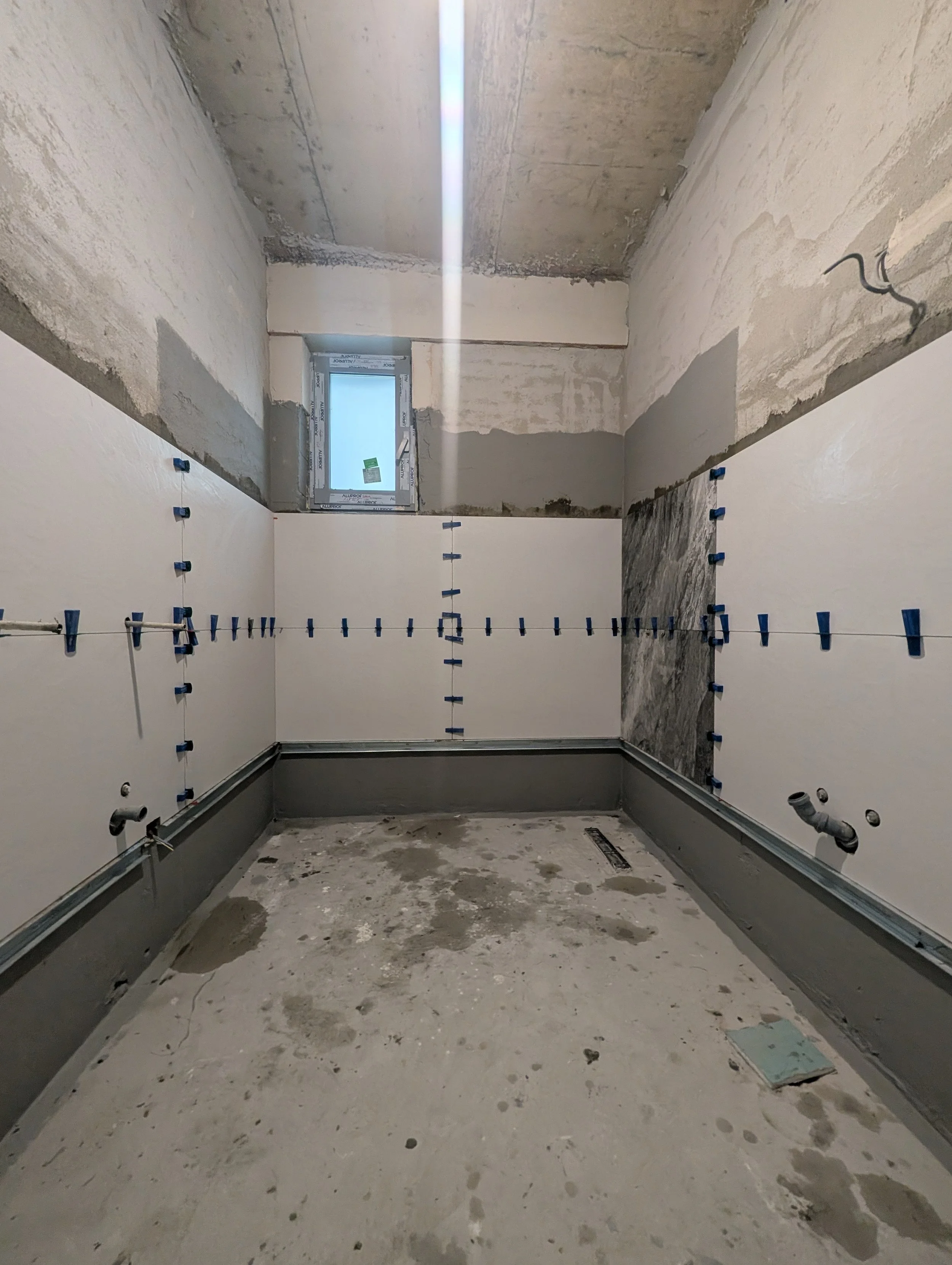 An unfinished bathroom under construction with partially tiled walls, plumbing pipes visible, and construction debris on the floor.