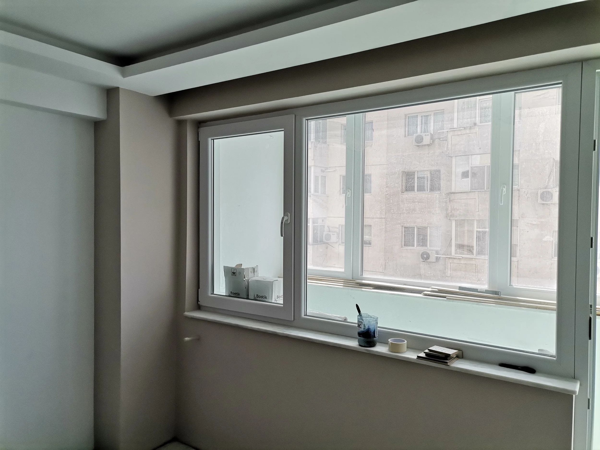 A room with a large window, partially open, revealing a cityscape. The windowsill has a few objects, including a black container, a roll of tape, a small box, and some papers. The room appears to be under renovation or decoration, with boxes labeled 