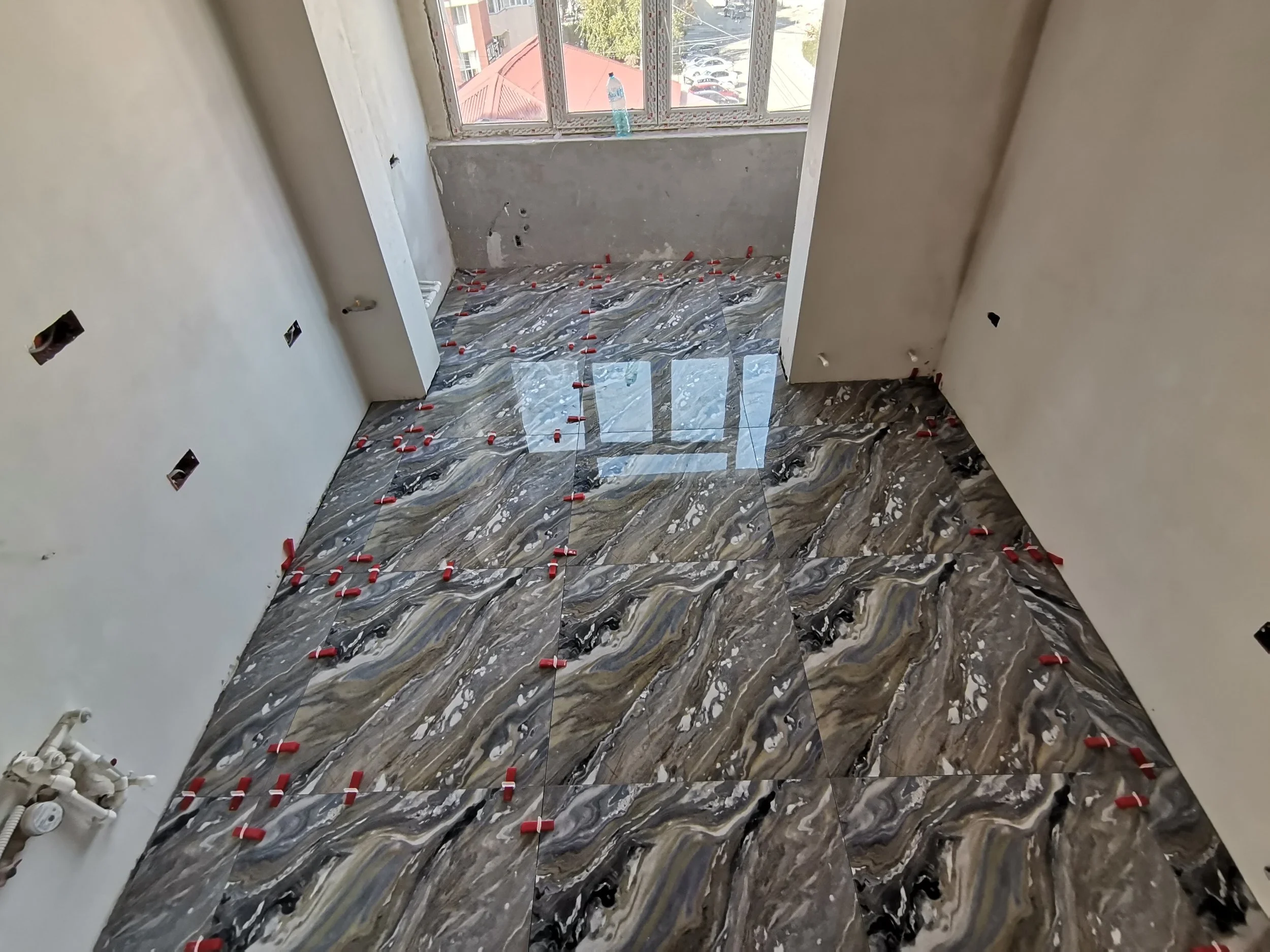 The image shows a room under construction with tiled flooring that has a marbled pattern of black, brown, and gray. There are small red spacers placed between the tiles for grout. The walls are unfinished with electrical outlets and pipes visible. A 