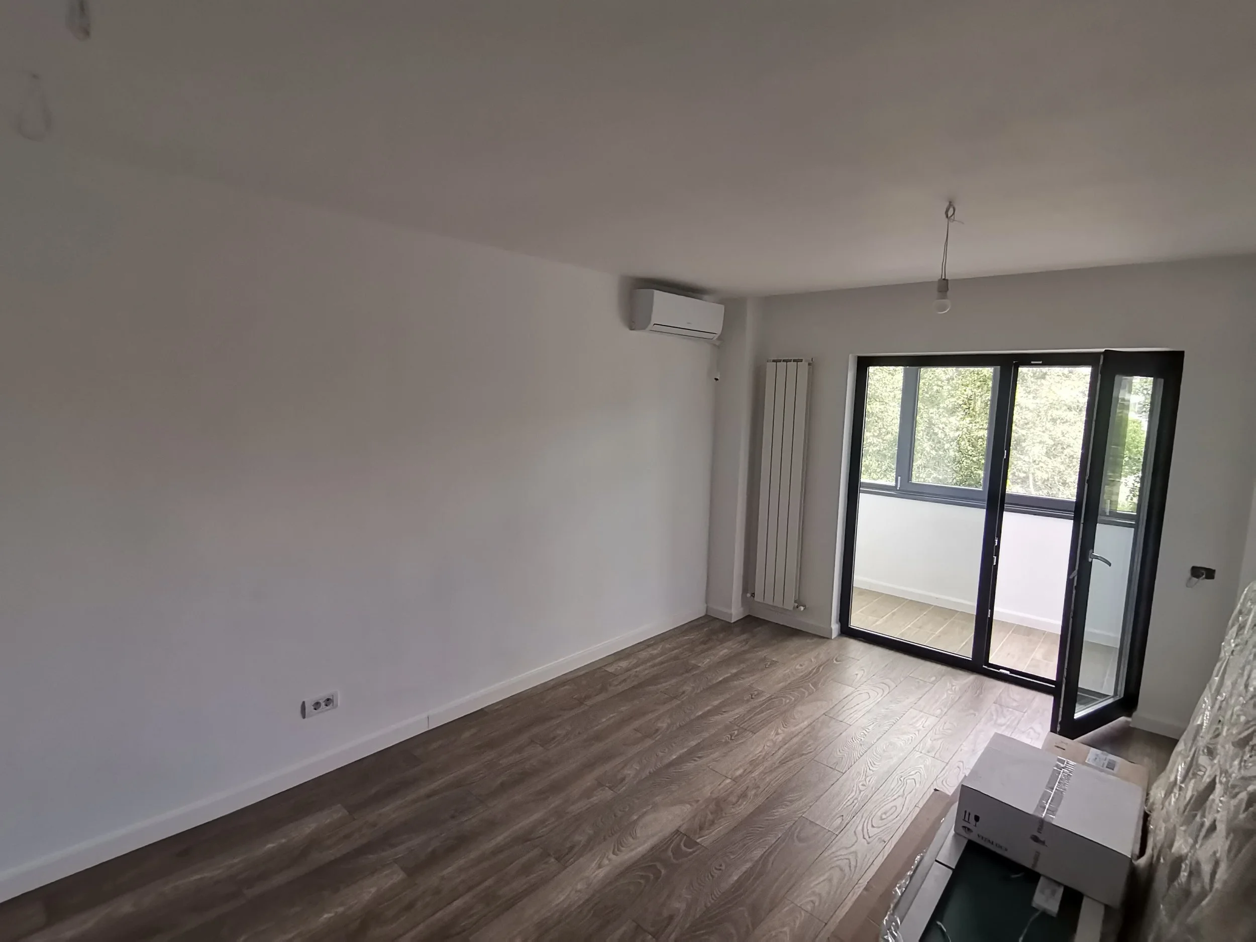 A modern, unfurnished room with white walls, wood flooring, a black-framed glass door leading to a balcony, an air conditioning unit, a radiator, and an electrical outlet.