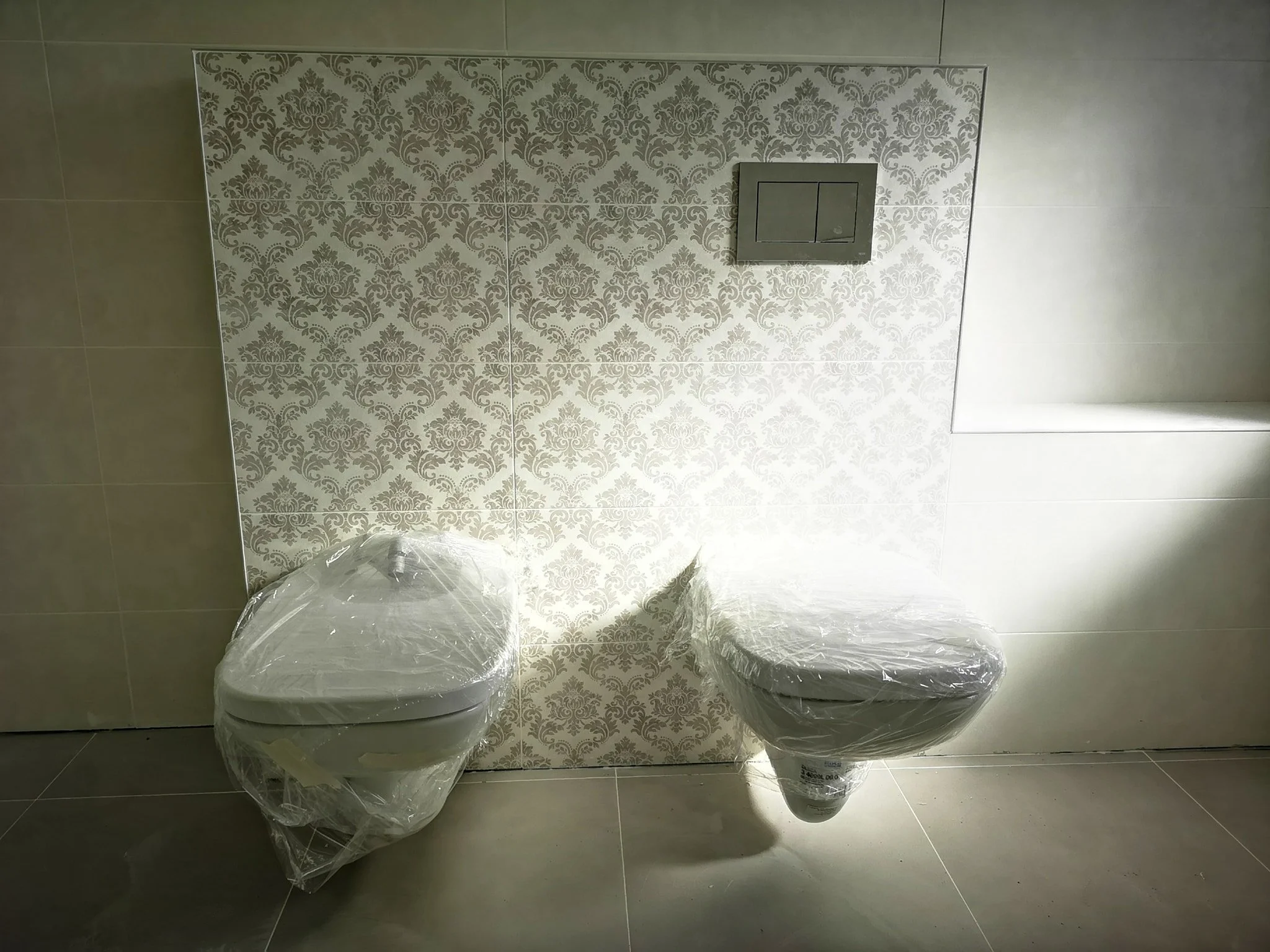 Two newly installed toilets wrapped in plastic, set against a decorative damask-patterned wall in a bathroom.