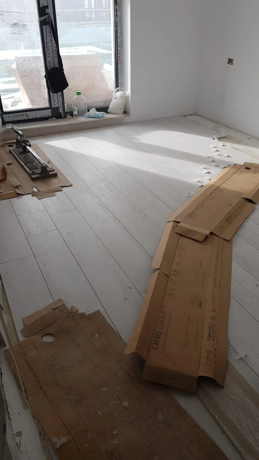 Room under construction with white flooring, large window with sunlight, and construction tools and materials scattered around.