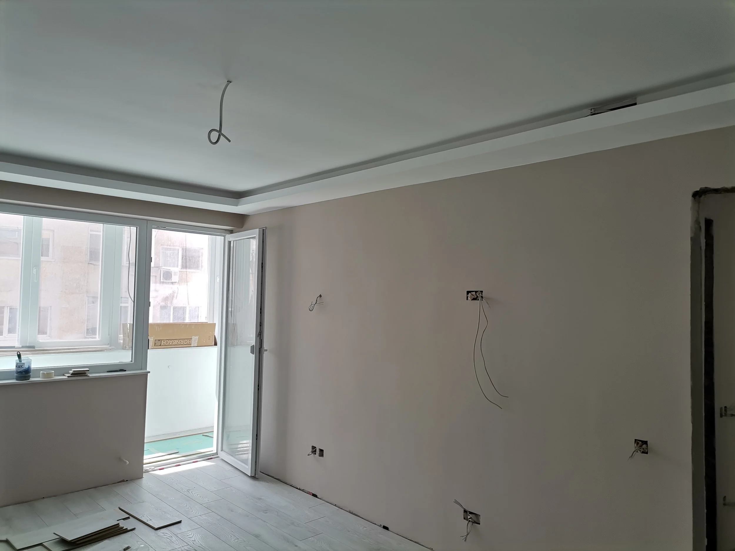 Interior of a room under construction with a ceiling cable hanging, a window with multiple glass panes, and construction materials on the floor.