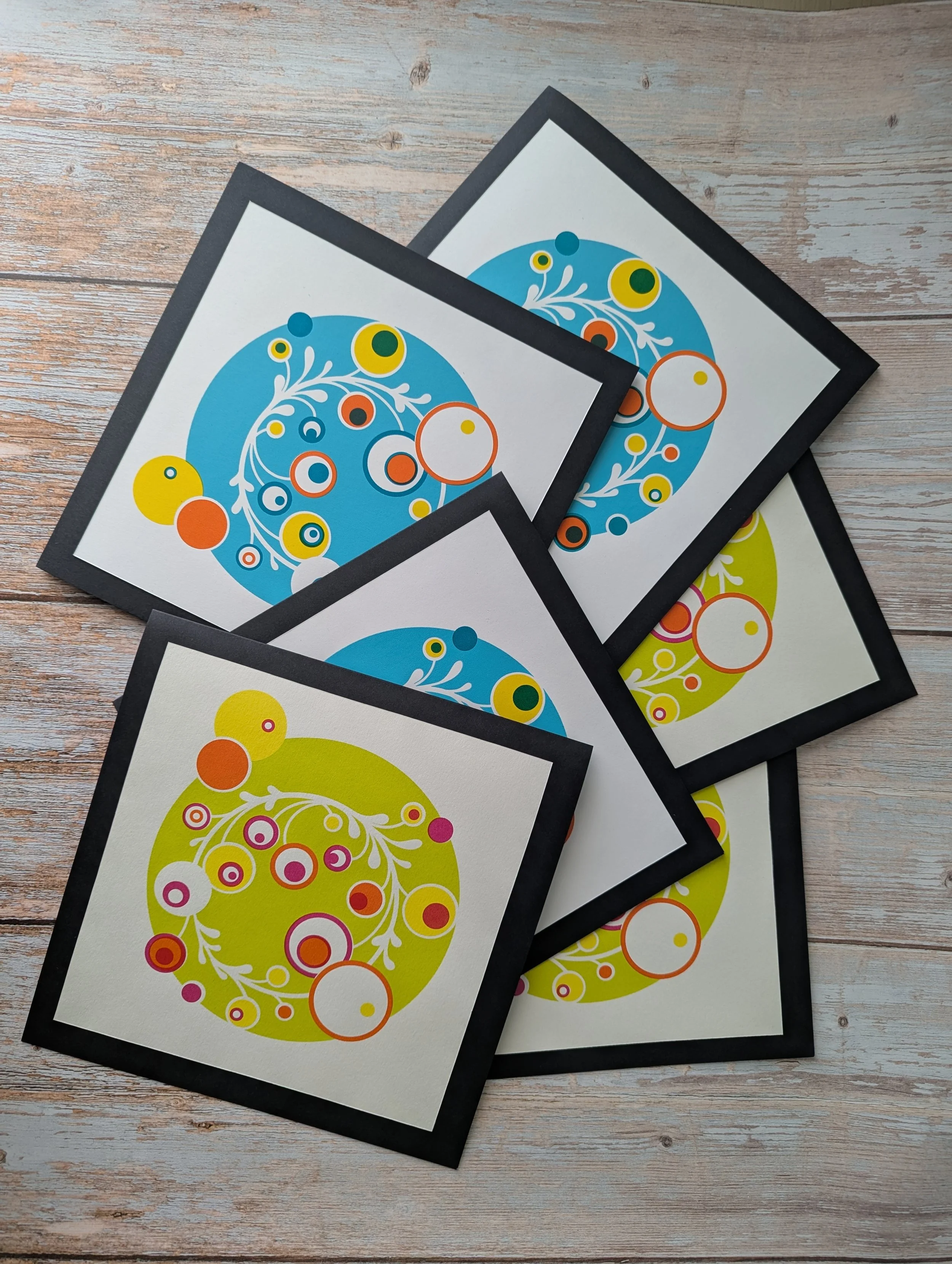 Multiple colorful abstract circular designs with white floral patterns on white backgrounds, framed in black, spread out on a wooden surface.