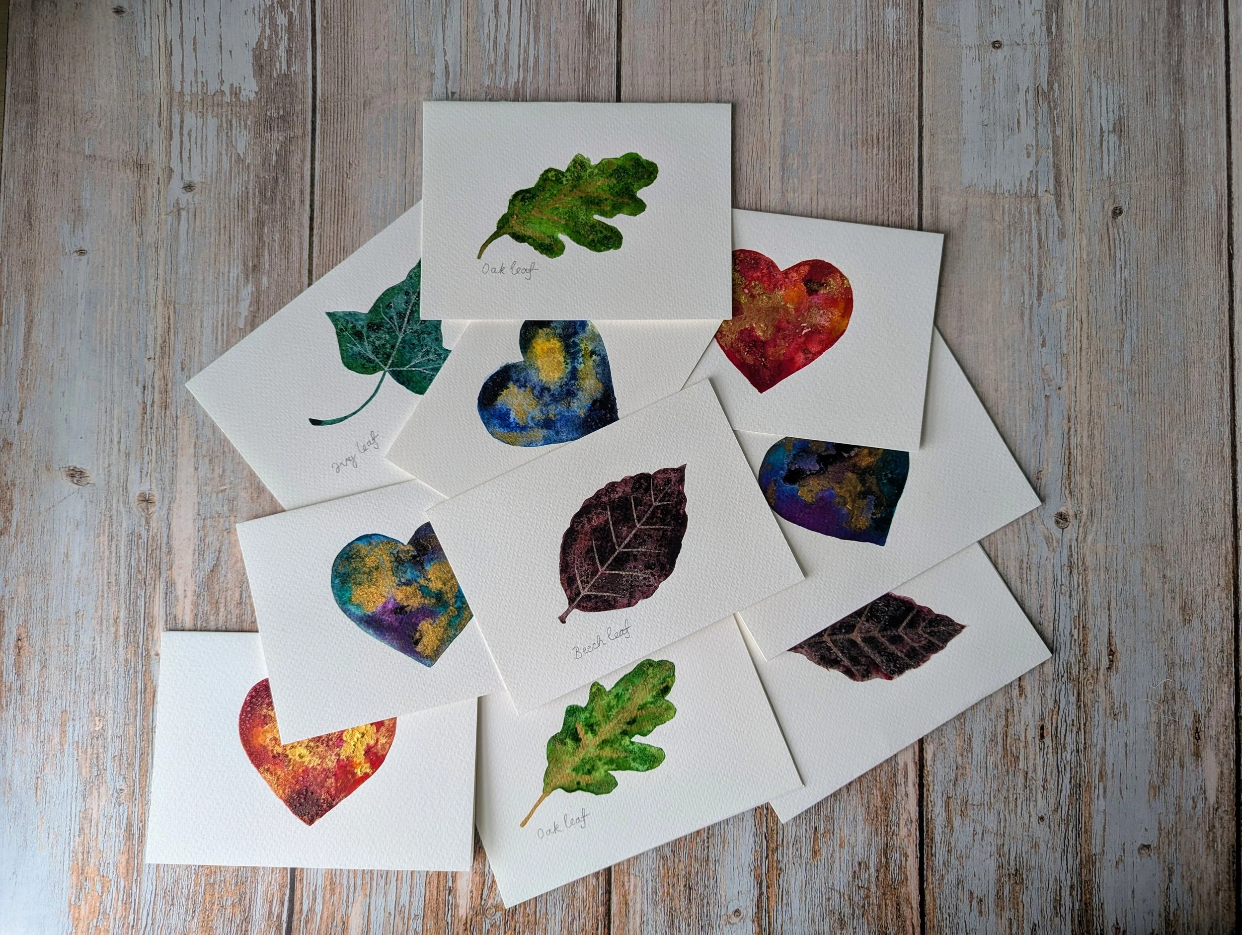 A selection hand painted cards, decorated with watercolor leaves and heart illustrations on textured white paper, scattered on wooden surface.