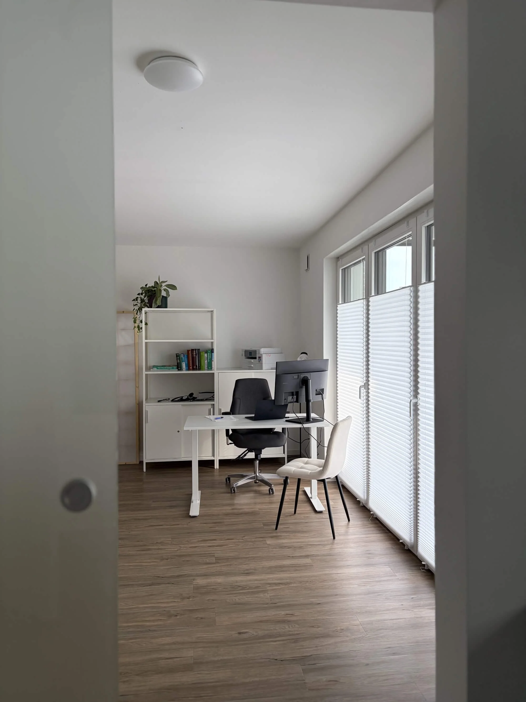 Ein Blick in ein helles und modernes Sprechzimmer mit einem Schreibtisch, bei dem ein Drehstuhl auf der einen, ein Besucherstuhl auf der anderen Seite steht, mit einem Regal mit Büchern und Pflanzen und großen Fenstern mit Jalousien.