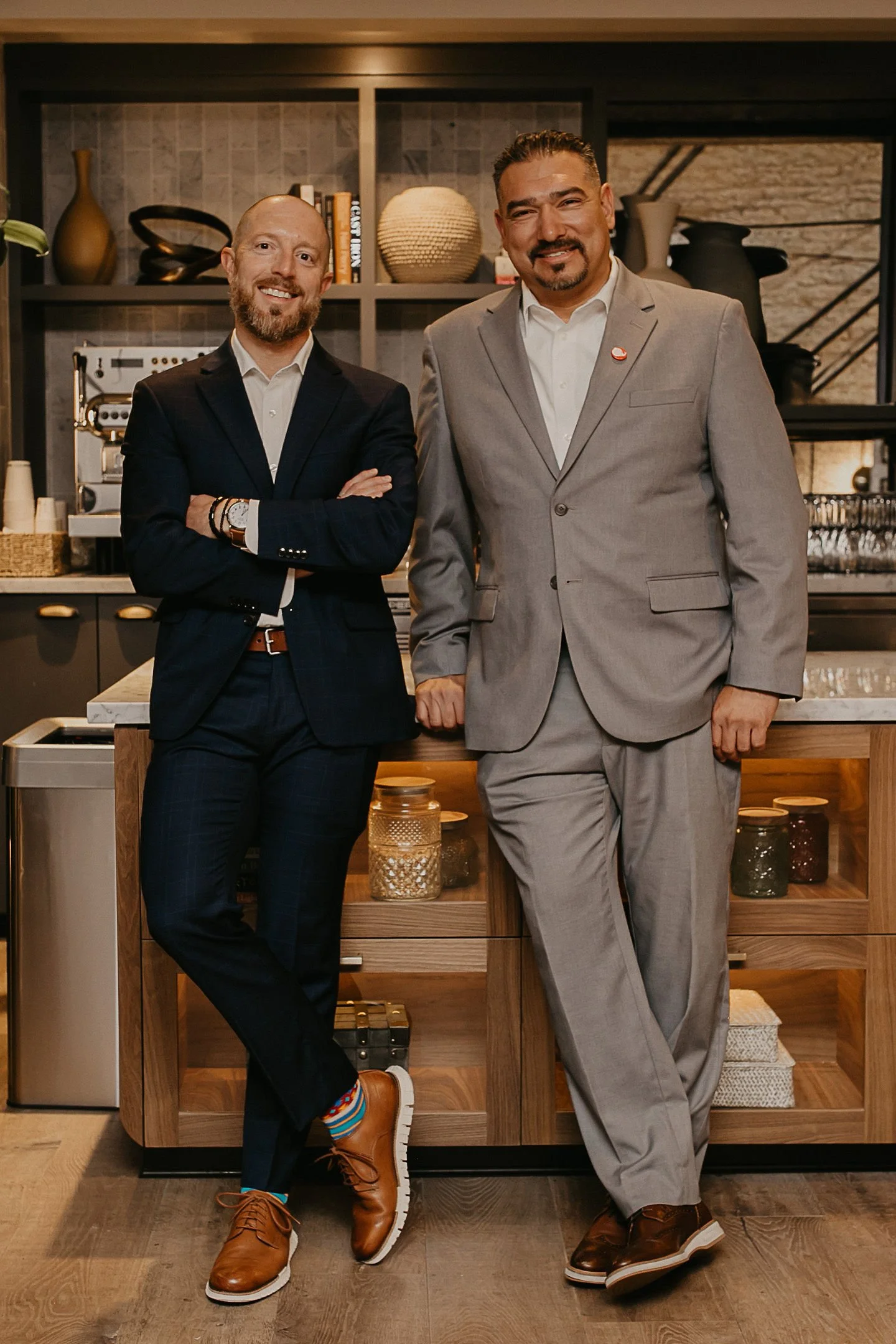 Two men in suits standing in a modern kitchen, smiling at the camera. The man on the left has a beard, is wearing a dark suit with a white shirt, colorful socks, and brown shoes. The man on the right has short dark hair, a goatee, wears a light gray suit with a white shirt, and brown shoes. The kitchen features open shelves with decorative items, books, and a coffee machine.