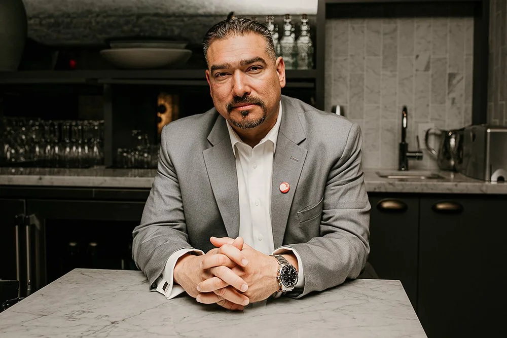 Man in a gray suit sitting at a marble table in a modern kitchen.