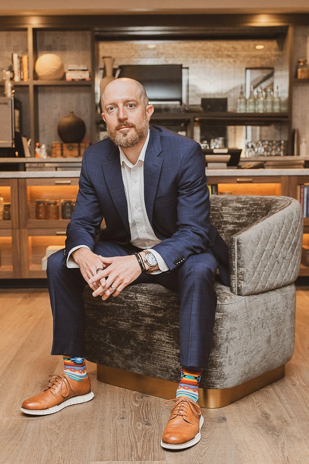 A man in a blue plaid suit with a white shirt, sitting on a modern gray armchair in a cozy, well-decorated living room or office space, wearing colorful patterned socks and tan shoes.