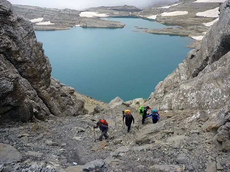 Grupo de esquiadores de montaña subiendo una pendiente rocoso con un lago de fondo.