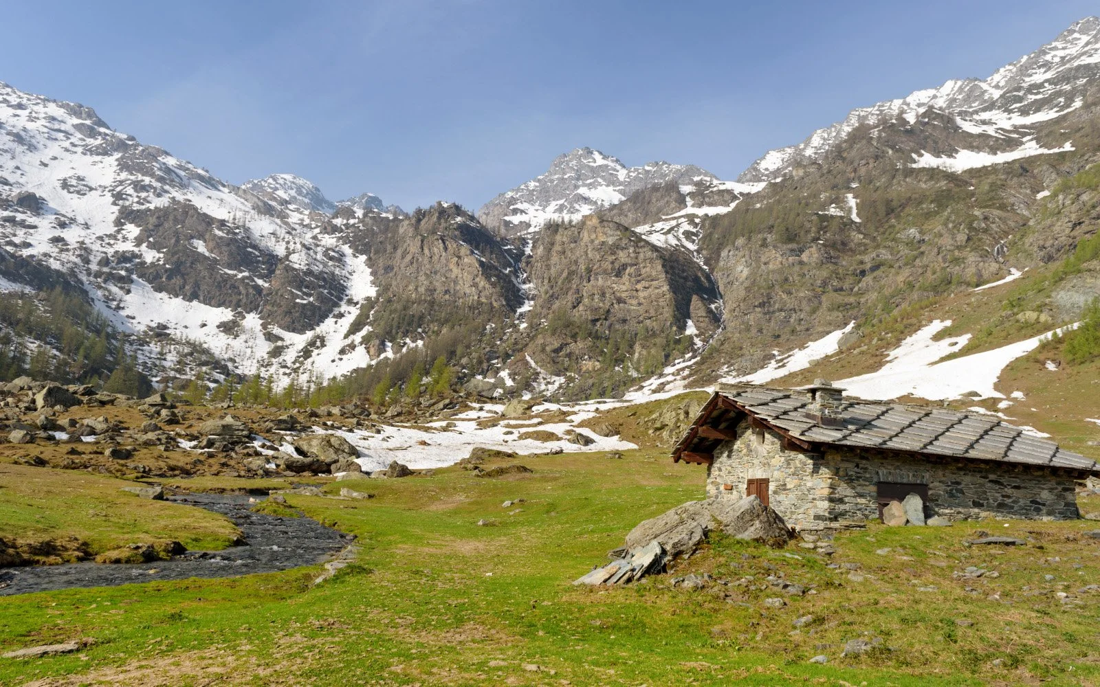 Casa de piedra en valle montañoso con nieve en las cumbres, río pequeño y césped verde.