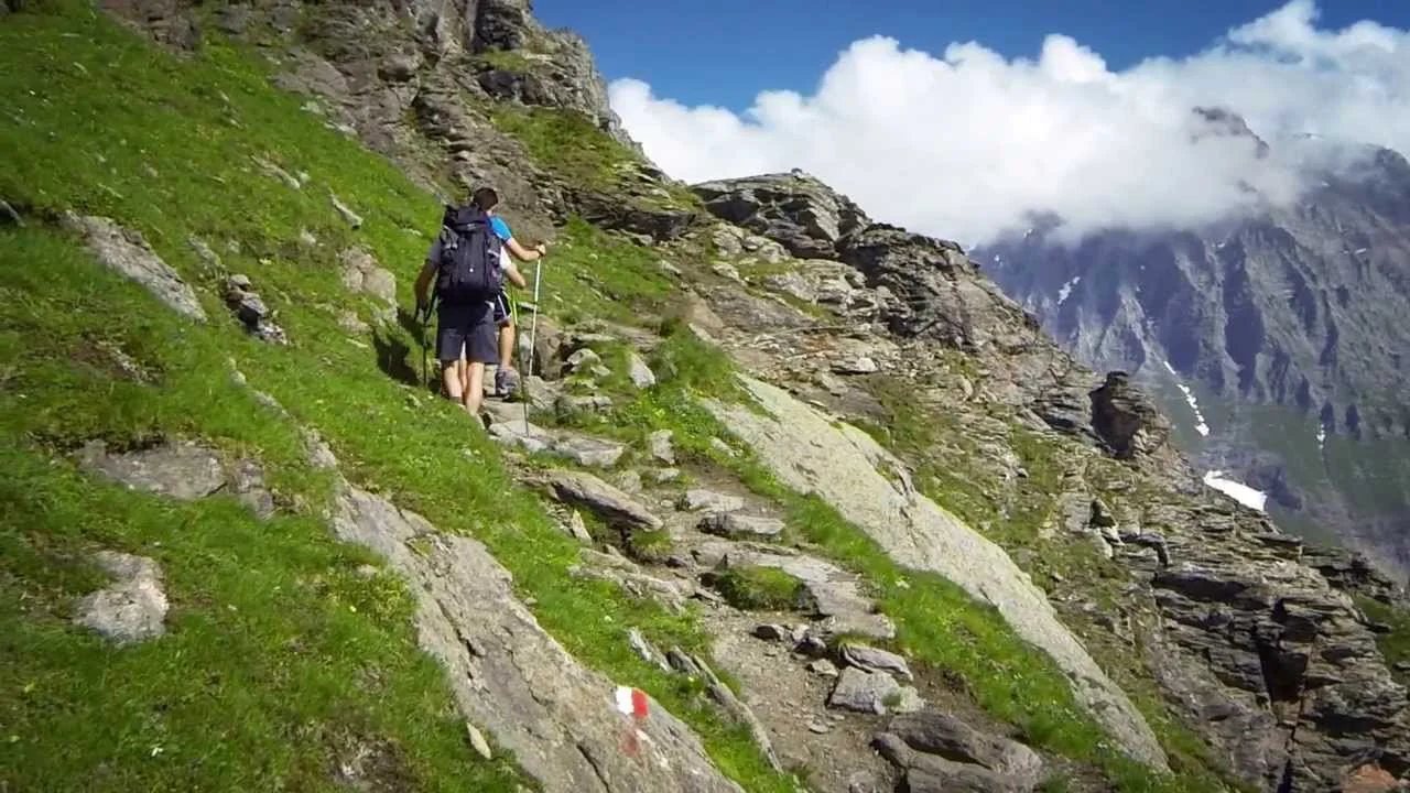 Dos personas haciendo senderismo en una ruta de montaña con vegetación verde y rocas, en un paisaje de montaña con nubes y picos nevados en el fondo.