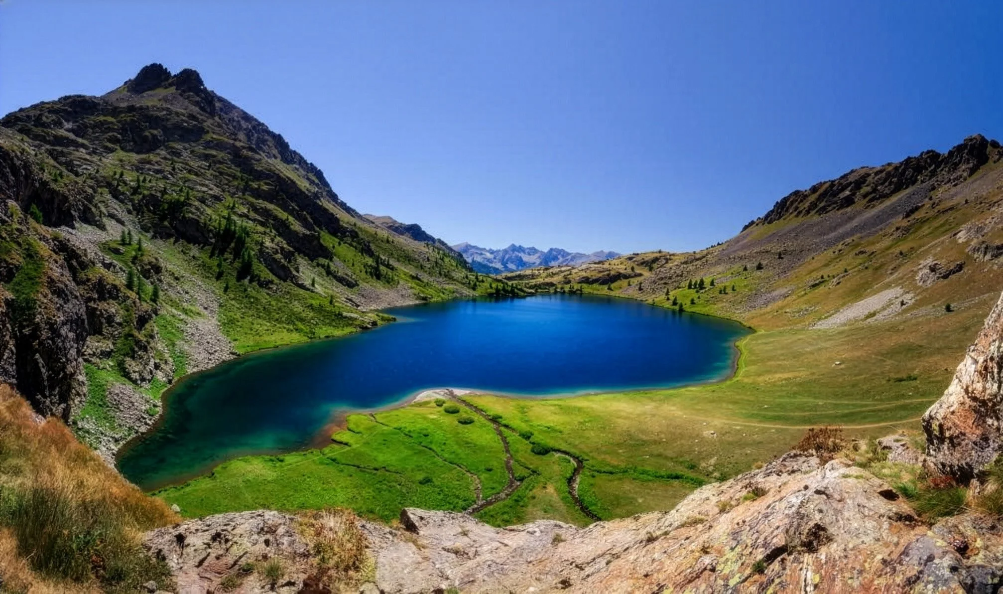 Lago en un valle rodeado de montañas con vegetación verde y rocas, bajo un cielo despejado.