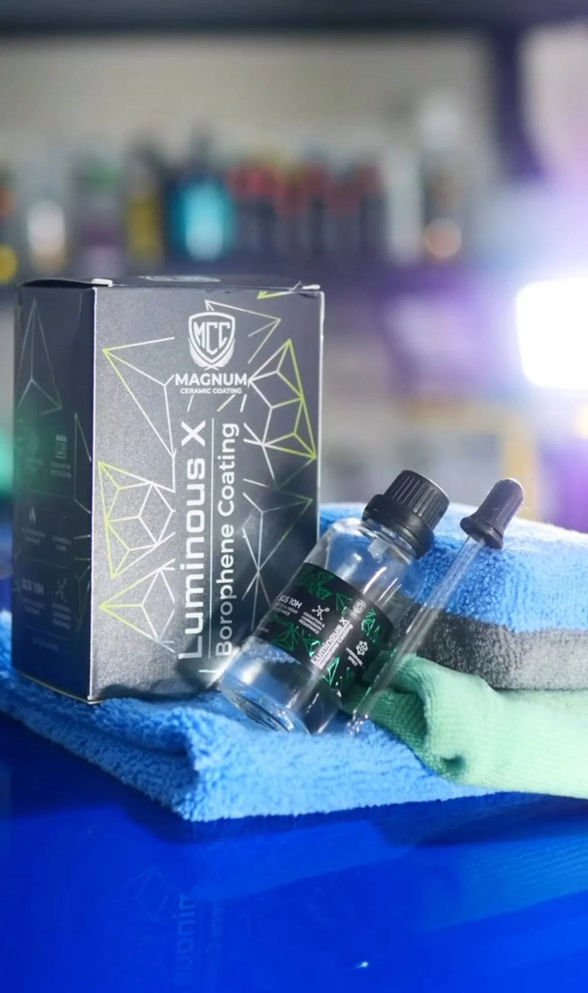 A box of Magnum ceramic coating, a small bottle of coating solution, and a microfiber cloth on a blue surface in a workshop setting.