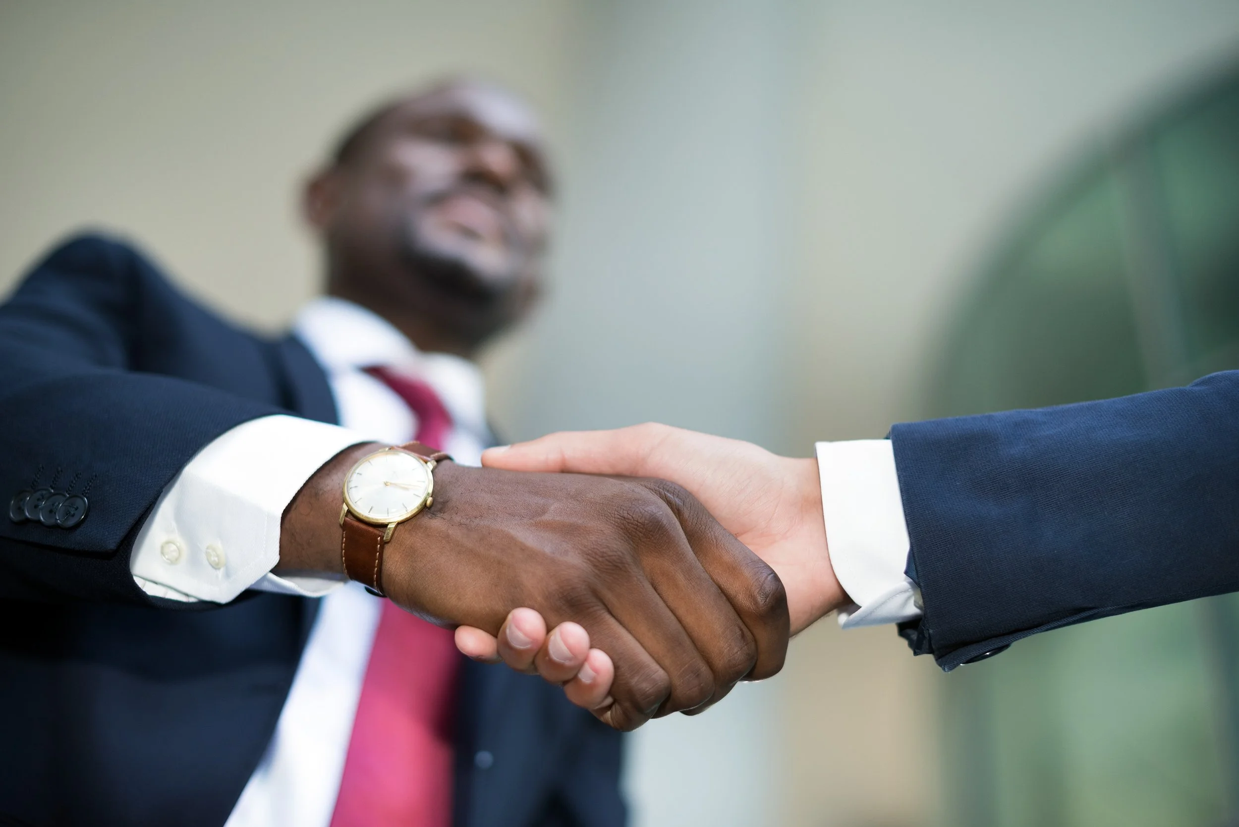 Close-up of two people shaking hands, one wearing a dark suit and a watch, and the other in a blue suit, with a blurred person in a suit and red tie in the background.