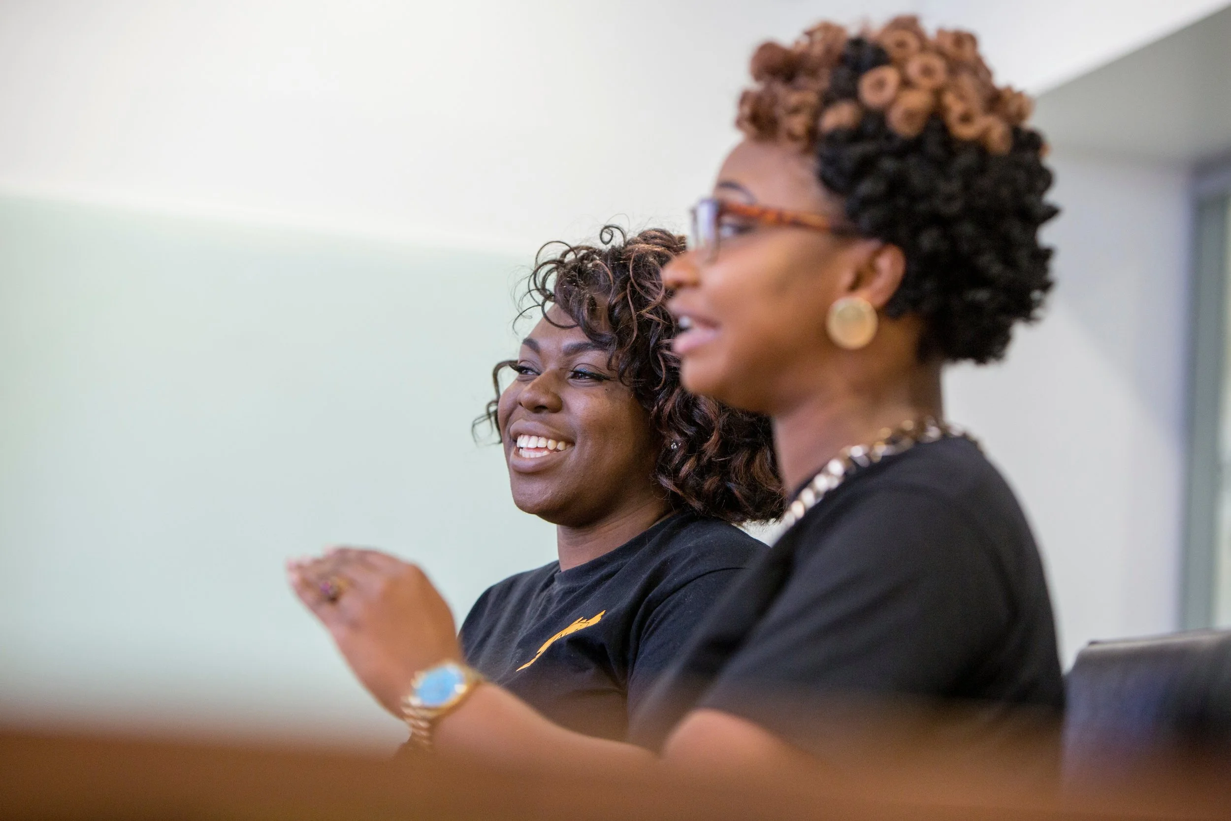 Two women with curly hairstyles and glasses smiling and engaging in a conversation or activity in an indoor setting.