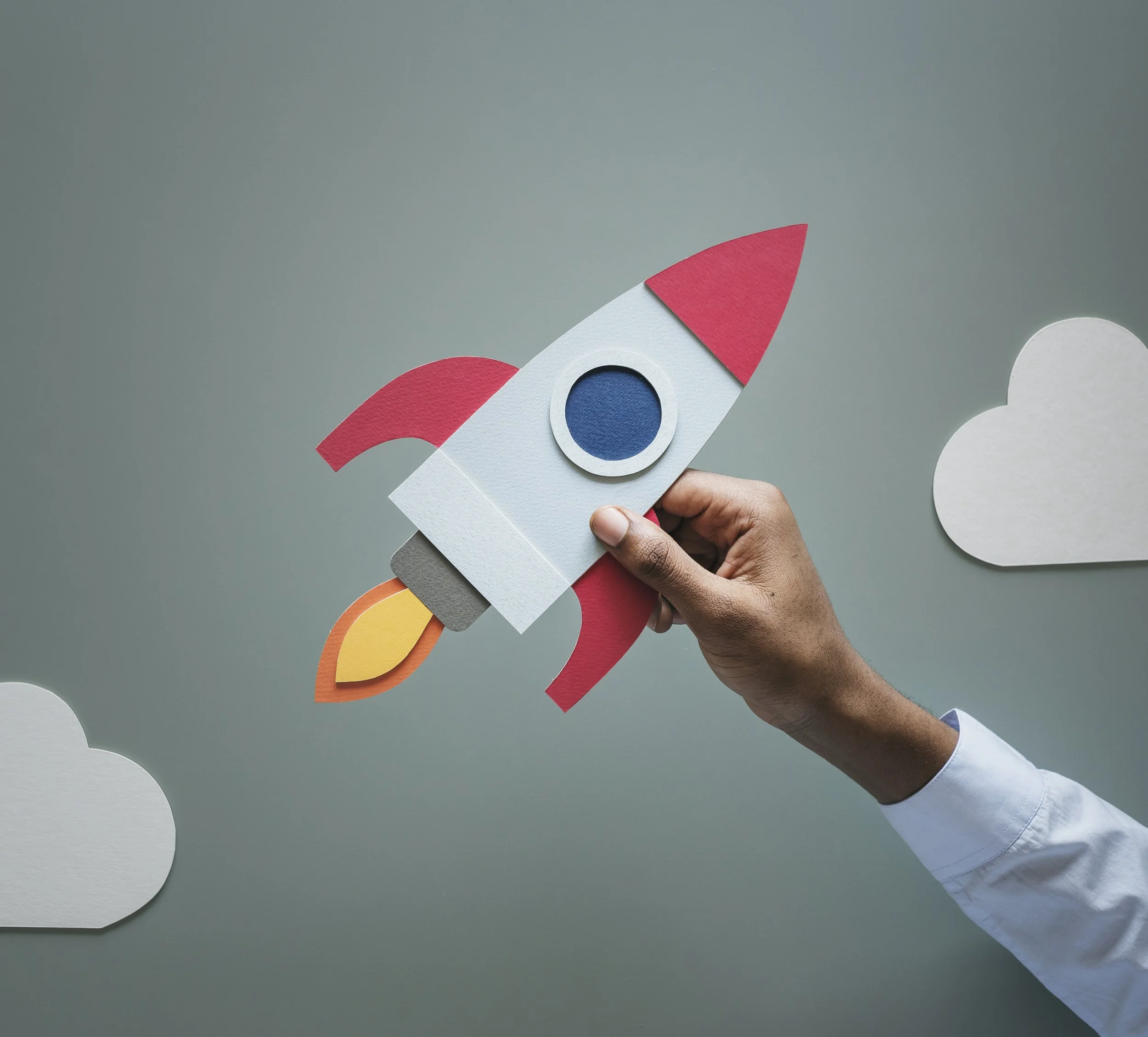 A person holding a paper cutout of a rainbow rocket in front of a plain gray background, with white cloud-shaped cutouts in the scene.