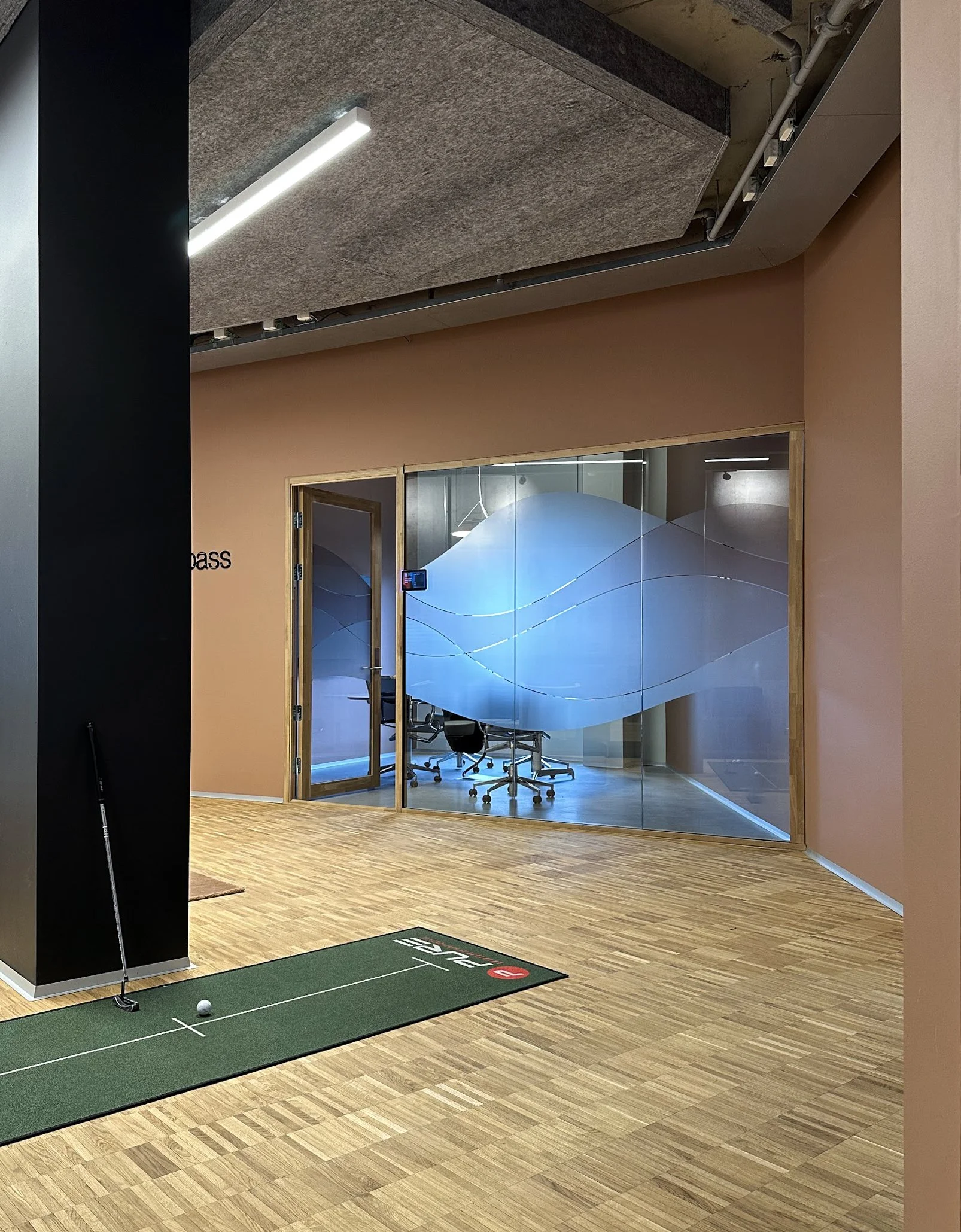 Sosial sone med en golfputtematta og en golfball i forgrunnen, samt glassvegg og konferanserom med stoler og bord bak.
