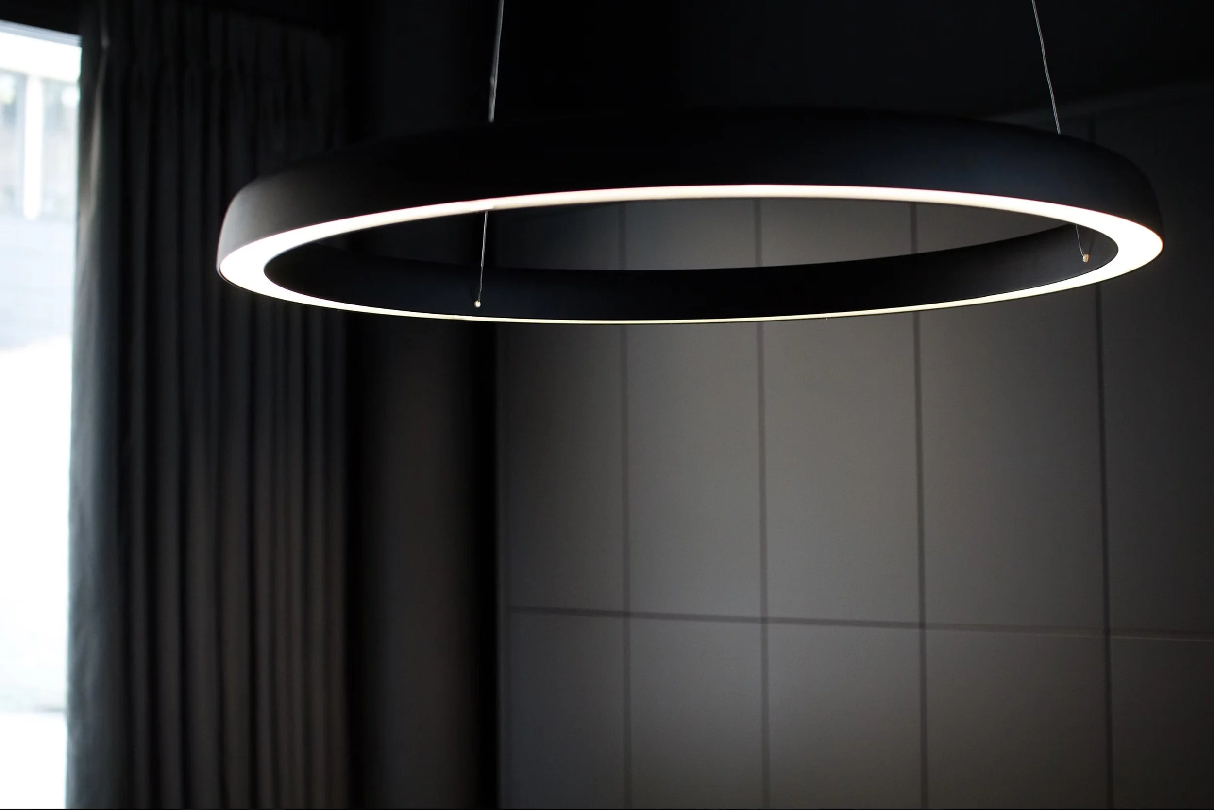 A black circular modern pendant light fixture hanging from the ceiling in a room with tiled walls. Natural light filters in from a window on the left side.