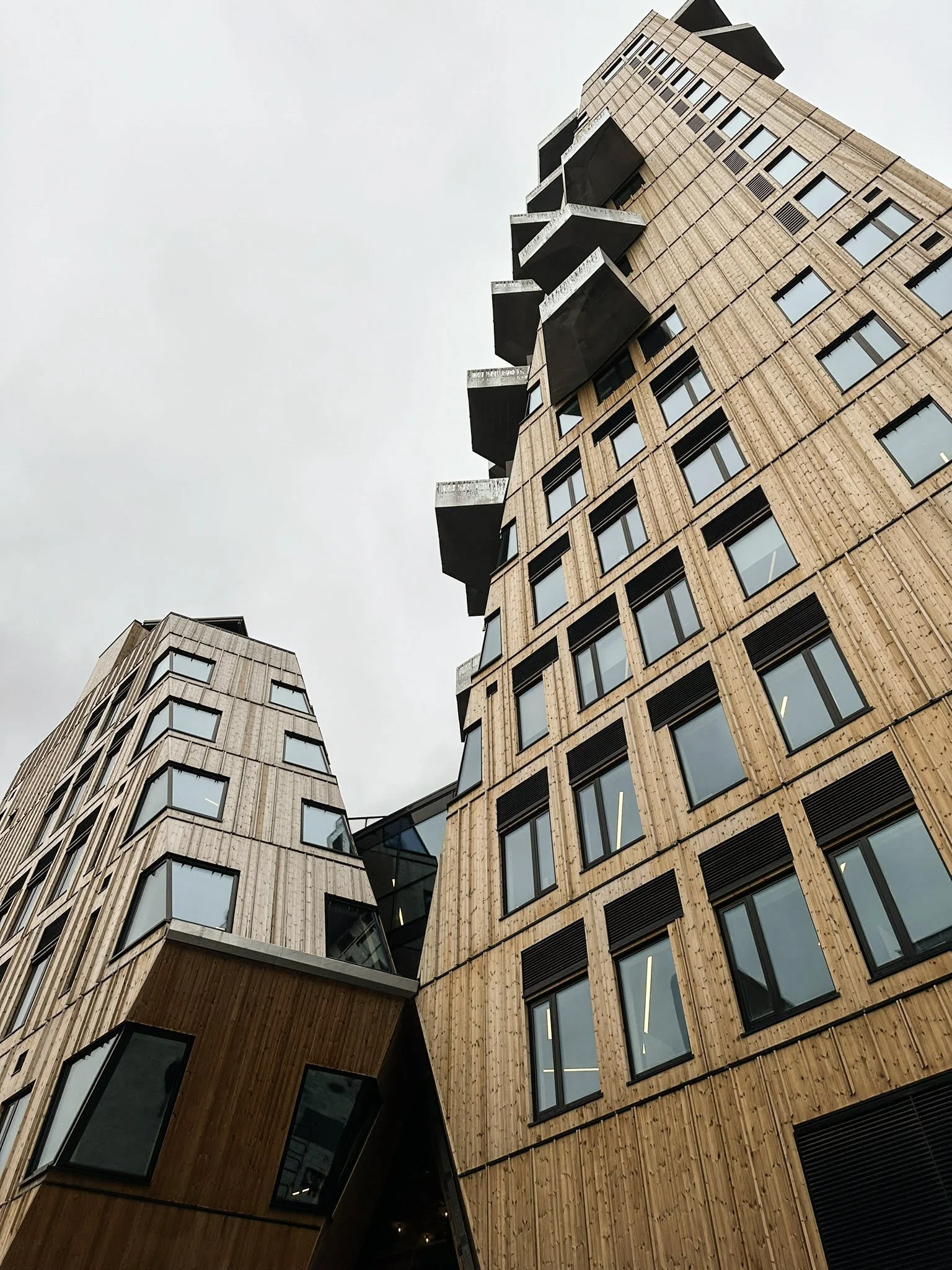 Fasade av moderne bygning med tre- og glassvinduer, og utstikkende balkonger, sett fra en lav vinkel mot en overskyet himmel.