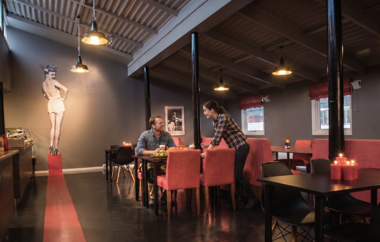 A man and woman sitting and talking at a table in a cozy restaurant, with a large black and white photo of a woman on the wall behind them, and candles on the tables. The restaurant has a modern style with pink chairs, black tables, and hanging lights.