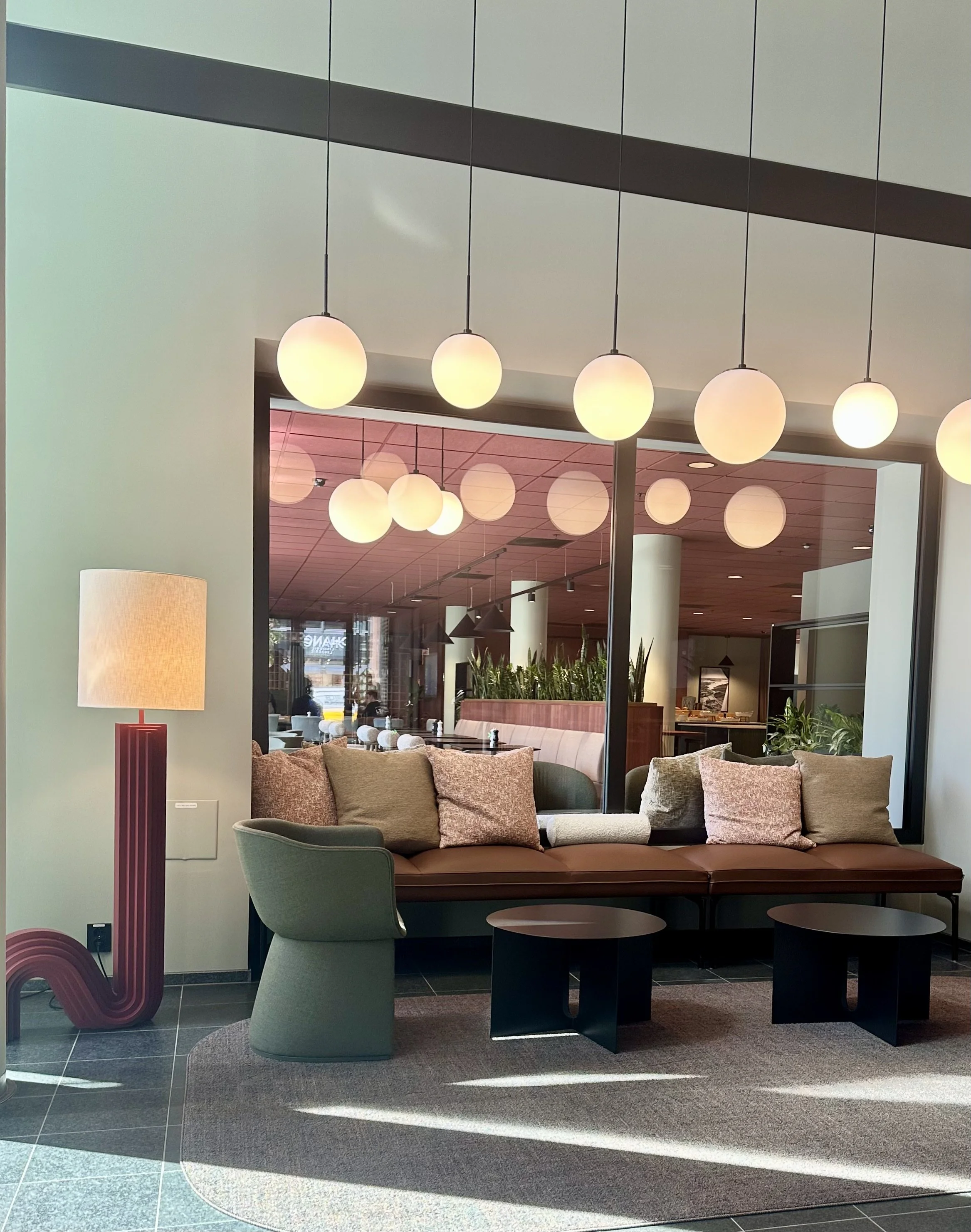 Liten lobby eller venteområde med sofa, to krakker, en gruppe med grønne planter, og en rekke av regulære, hvite, kulehengelamper som henger fra taket. En stor vindusvegg er synlig i bakgrunnen.