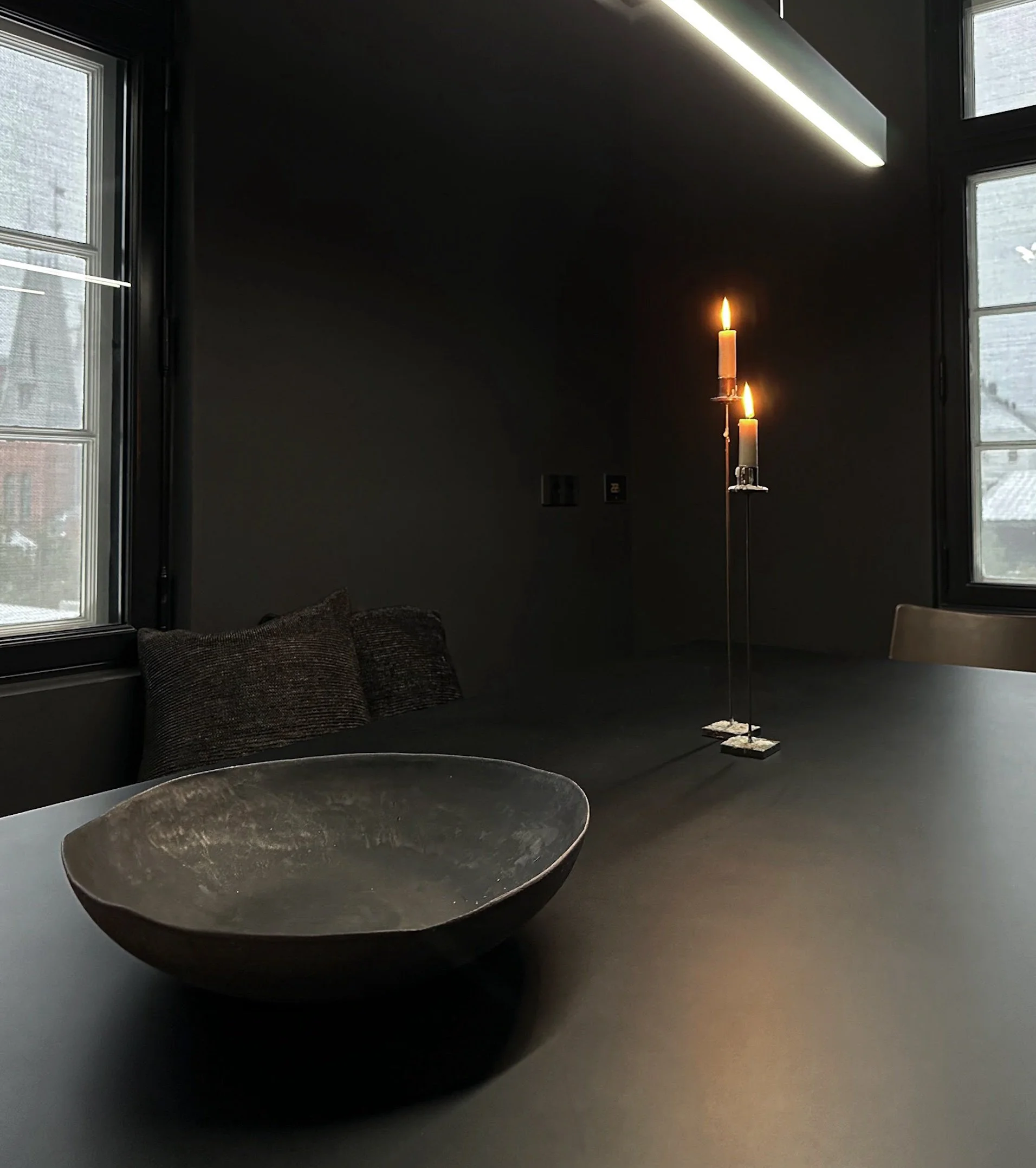 A round, dark-colored bowl on a table in a dimly lit room. The room has large windows with visible light coming through. There is a black object or furniture piece nearby, a few cushions, and a tall candlestick with two lit candles in the background. The candle flames are glowing against the dark wall.