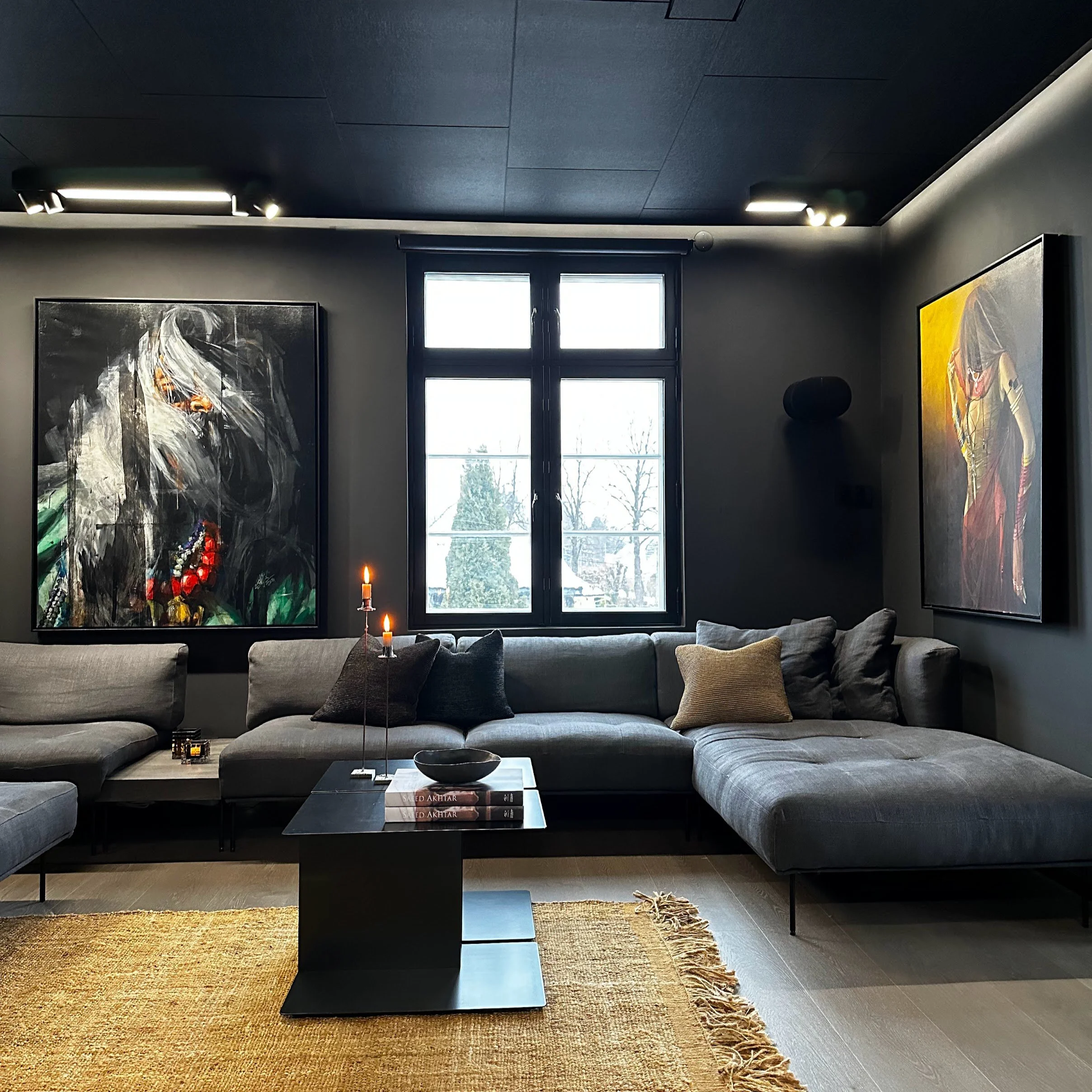 A modern living room with a large gray sectional sofa, a black coffee table with books and candles, abstract artwork on the walls, large windows, and a natural fiber rug.