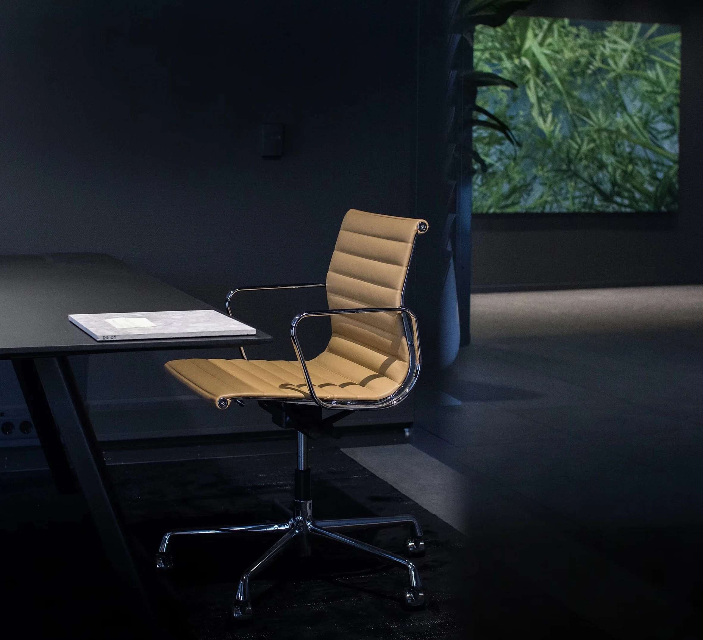 A modern conference room featuring a tan leather office chair with metal armrests and a black conference table with a light-colored pad or magazine on it. In the background, there is a wall with a large screen displaying an image of green foliage, and a small black object is mounted on the wall.