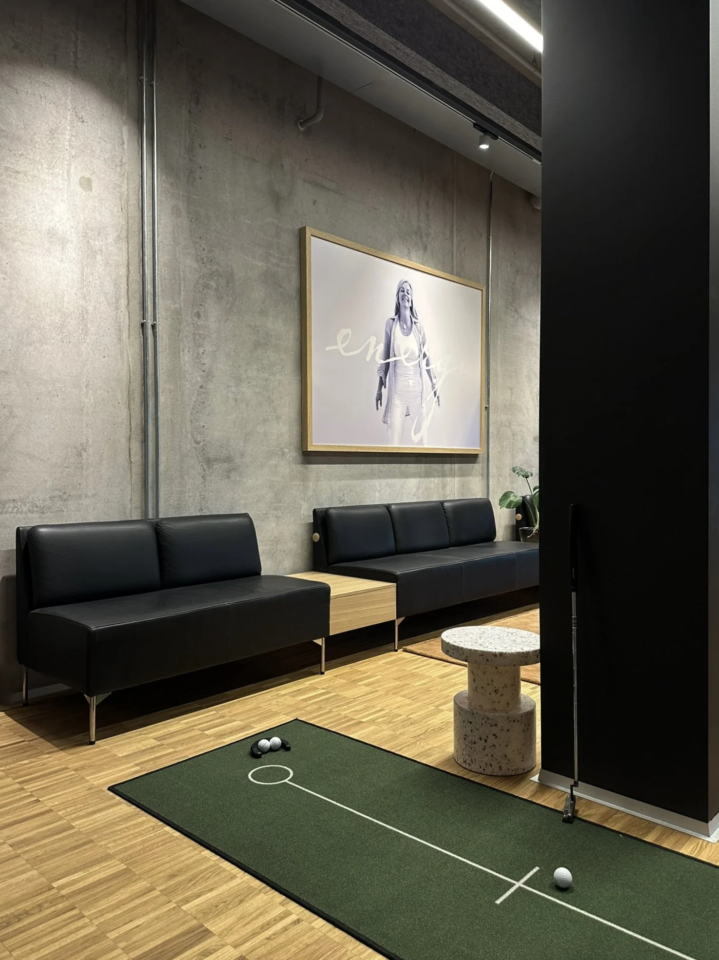 Innendørs loungeområde med svart sete, et lite bord, en daman golf putting matte, et bilde av en kvinne på veggen, og planter.