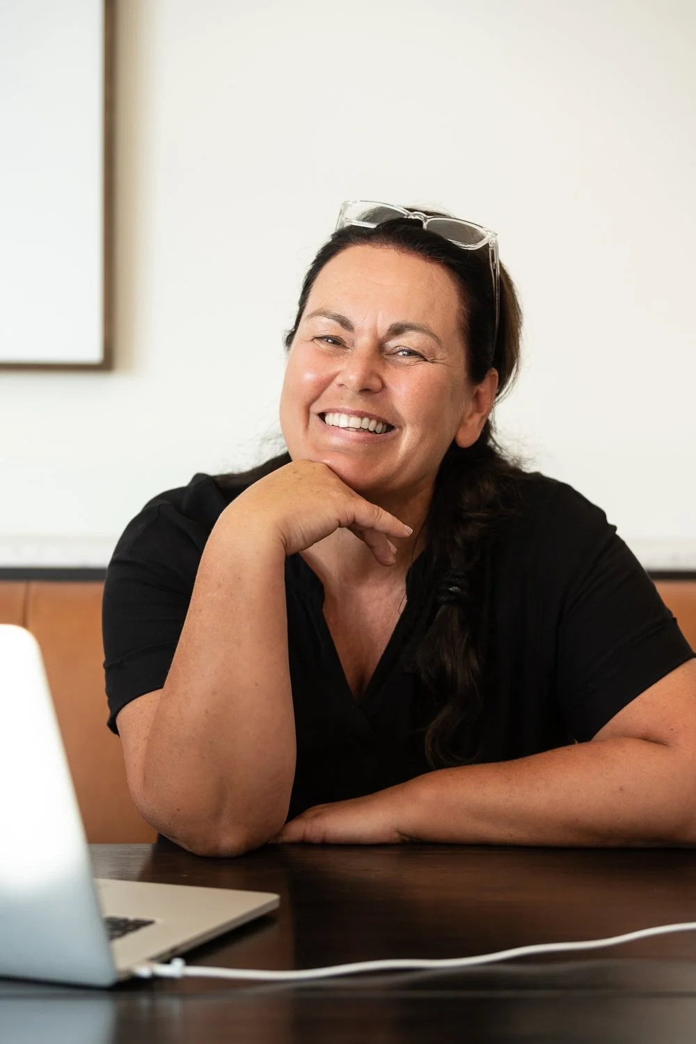 A middle-aged woman with dark hair, wearing a black shirt, smiling and resting her chin on her hand at a desk with a laptop in front of her.