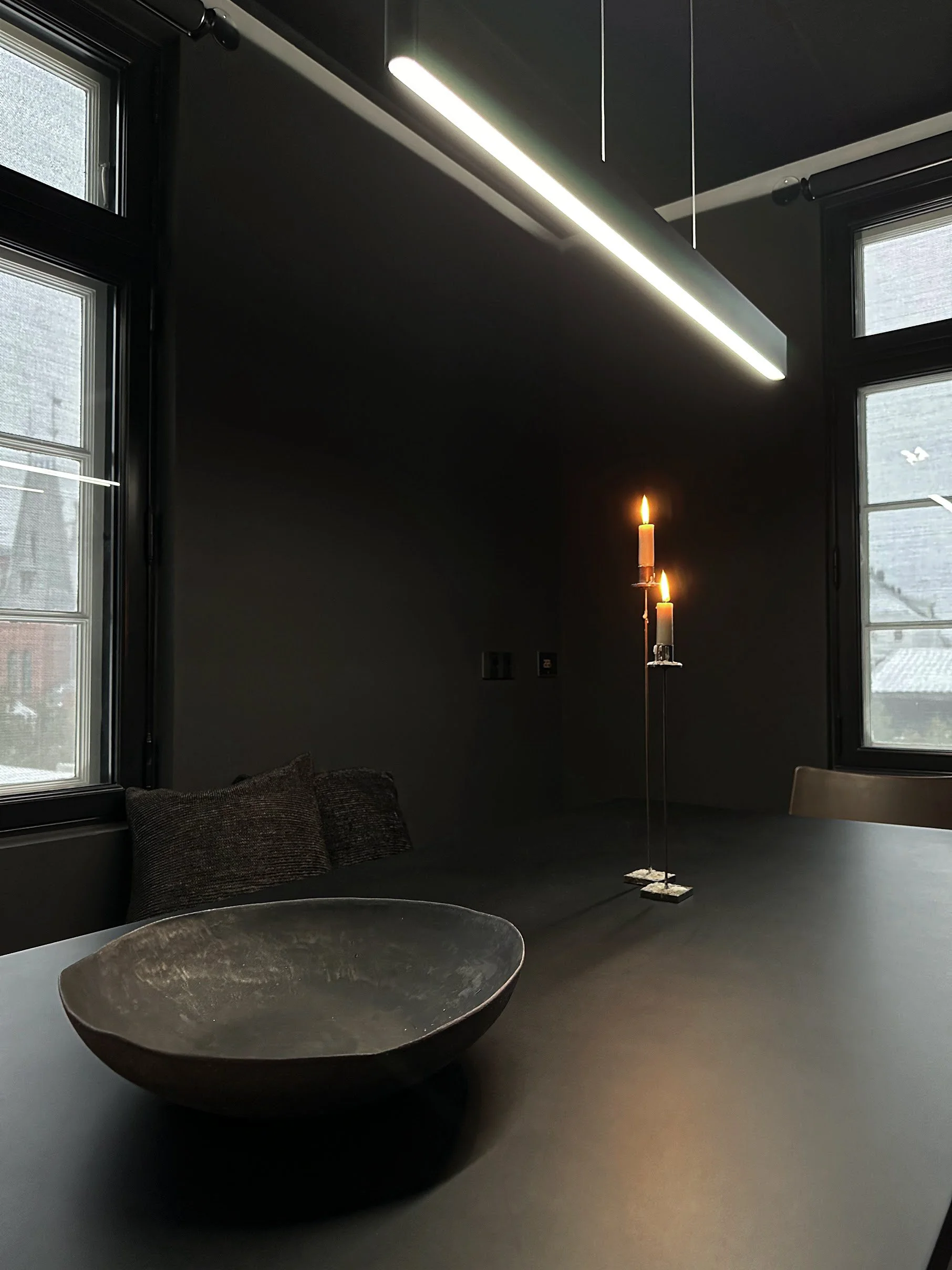 Modern interior room with black walls, large windows, a sleek black hanging light fixture, a tall candle holder with candles, a round bowl-shaped decorative object, and a cushioned seating area with pillows.