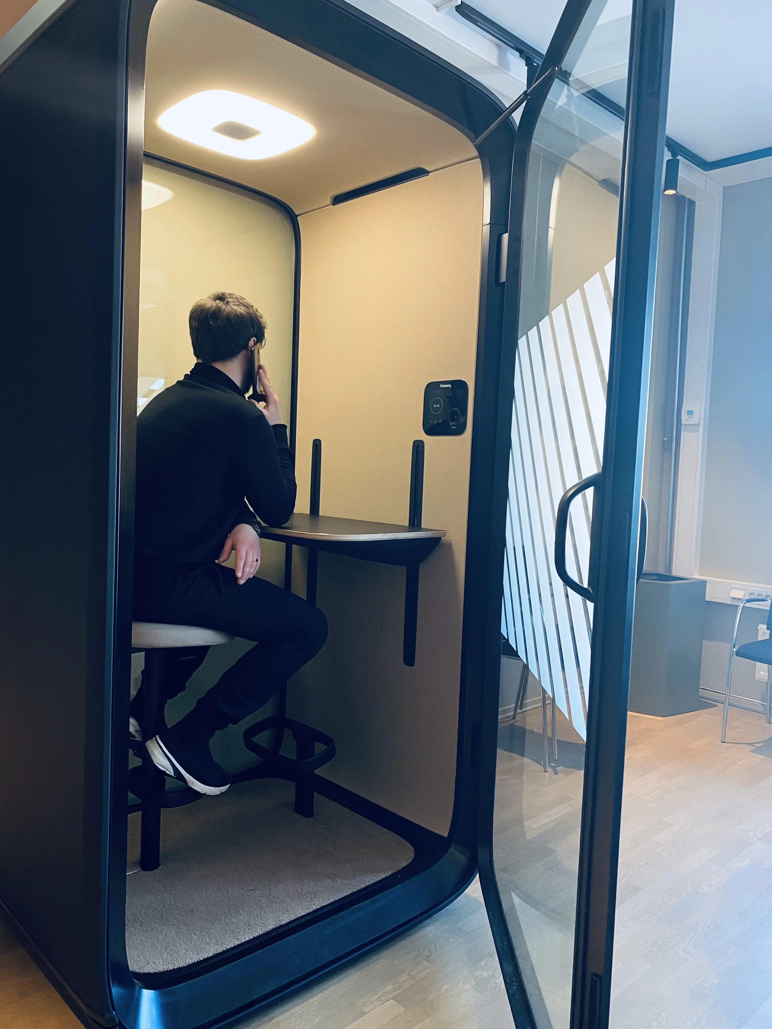 En person sitter i en lydisolert Phone Booth med beige innredning, snakker i telefonen, inne i et moderne kontor.