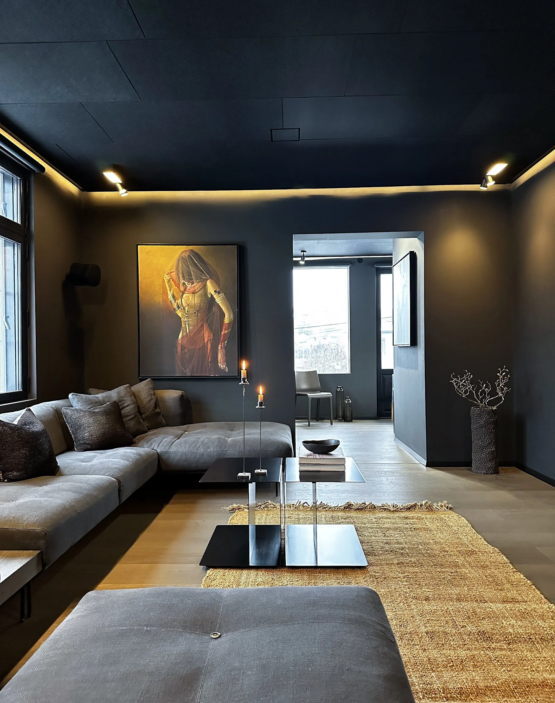 Modern living room with dark gray walls and ceiling, featuring a gray sectional sofa with cushions, a black rectangular coffee table with decorative items, a textured rug, a large framed artwork of a woman on the wall, and a corner vase with decorative branches. Multiple windows with blinds are visible, and ambient lighting highlights the room.