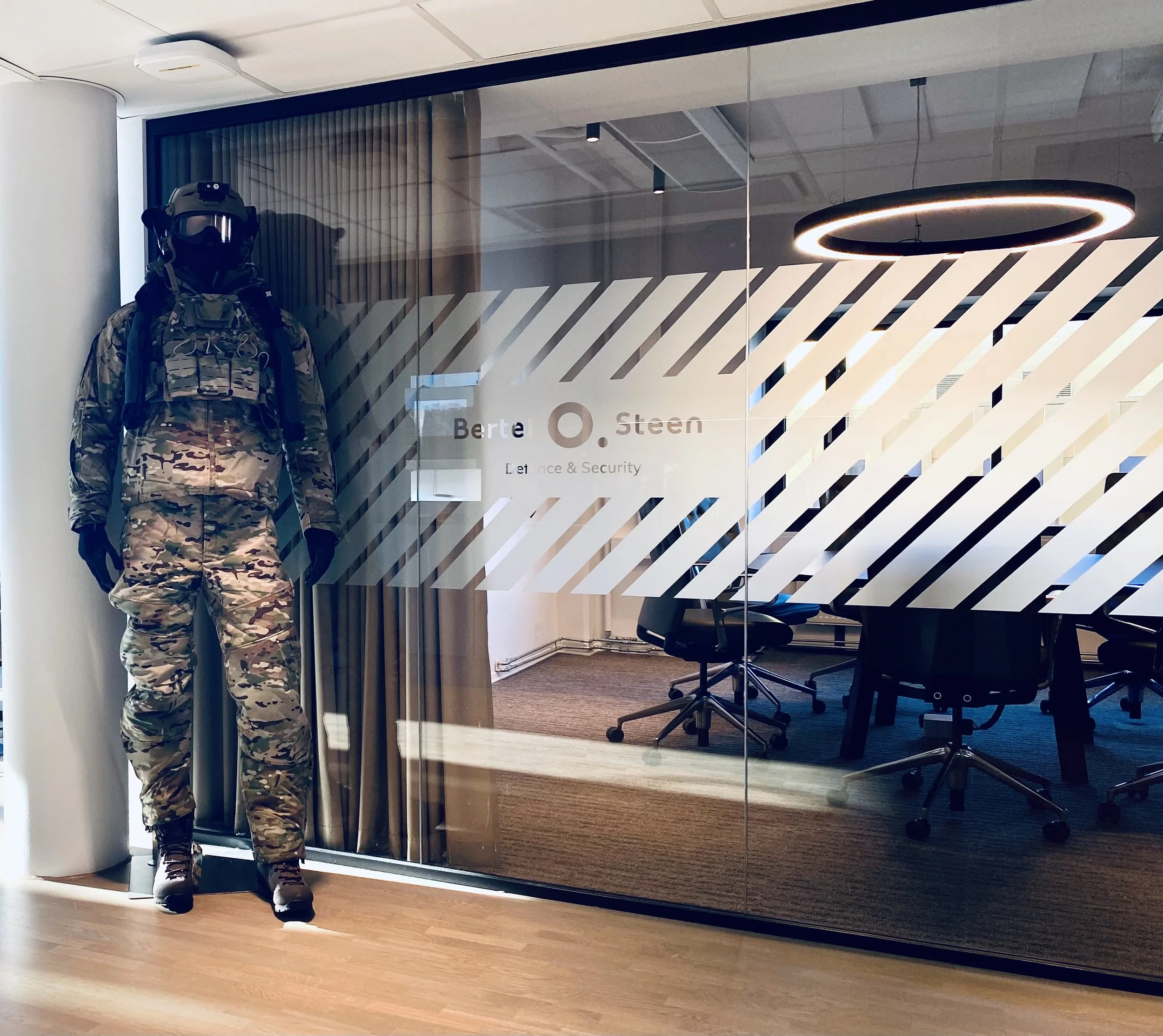 En person i militærtøy og hjelm står foran en glassvegg i et kontor med kontorutstyr og en rund taklampe.