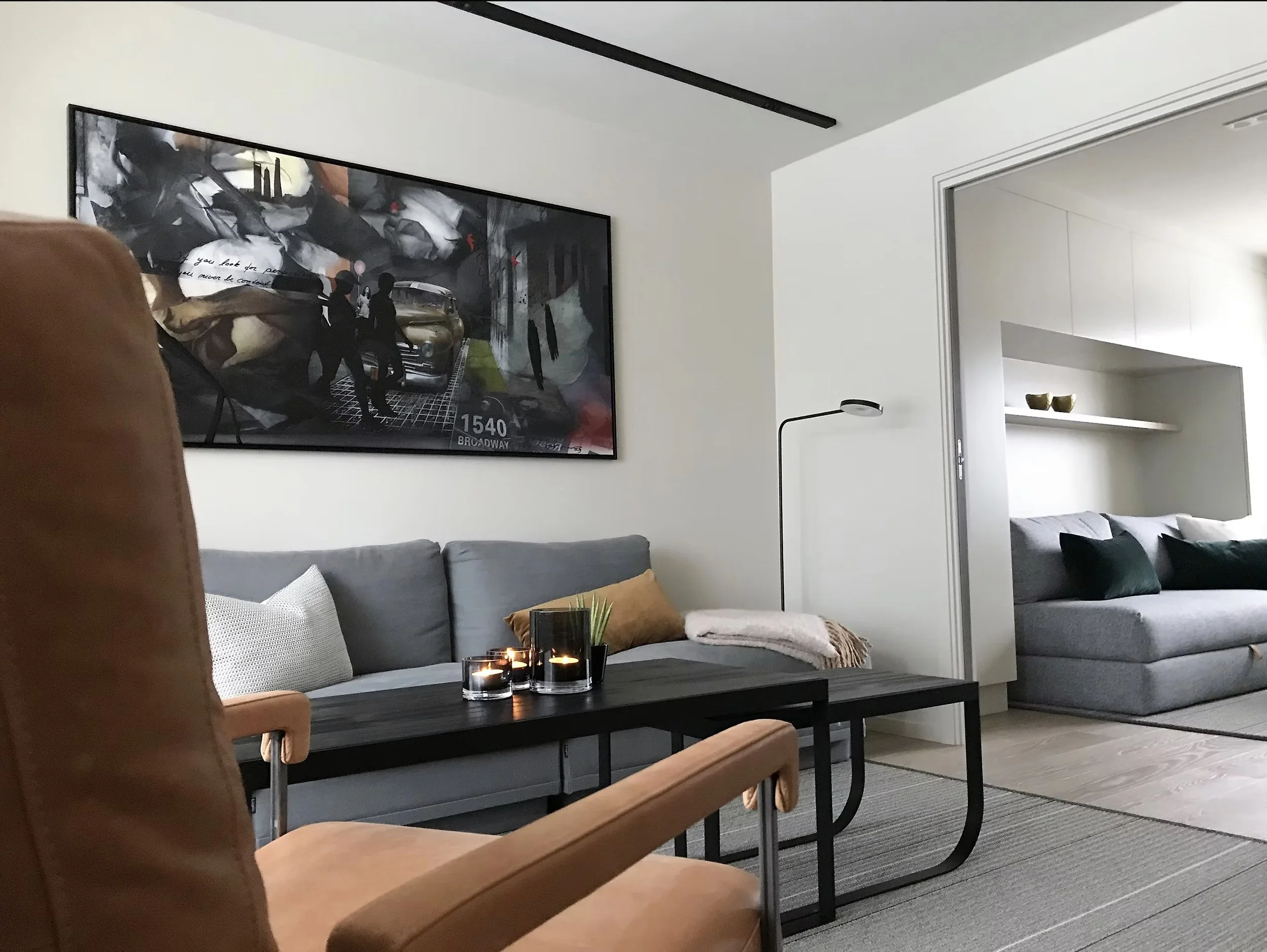 A living room with a gray sofa, a black coffee table with candles, a brown armchair, a large wall art, and a doorway leading to a bedroom with a bed and built-in shelves.