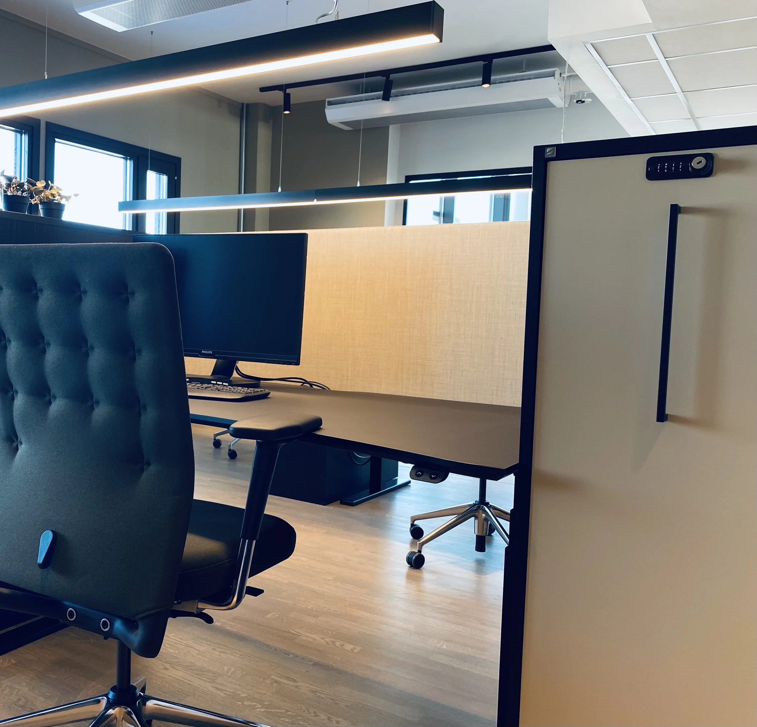 Et kontorarbeidsområde med dataskjerm, kontorstol og delte arbeidsplasser, med vinduer og taklys i bakgrunnen.