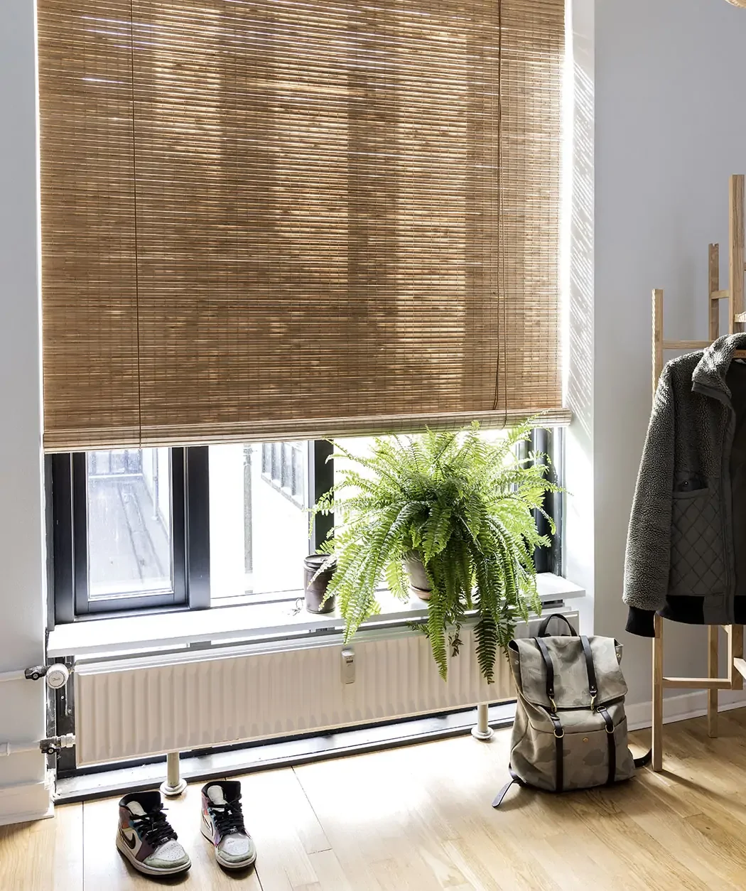 Innendørs bilde av et vindu med bambusrullgardin, en stor grønn fern plante på vinduskarmen, sandfargede sneakers på gulvet, og en ryggsekk og jakke ved siden av en klesstativ.