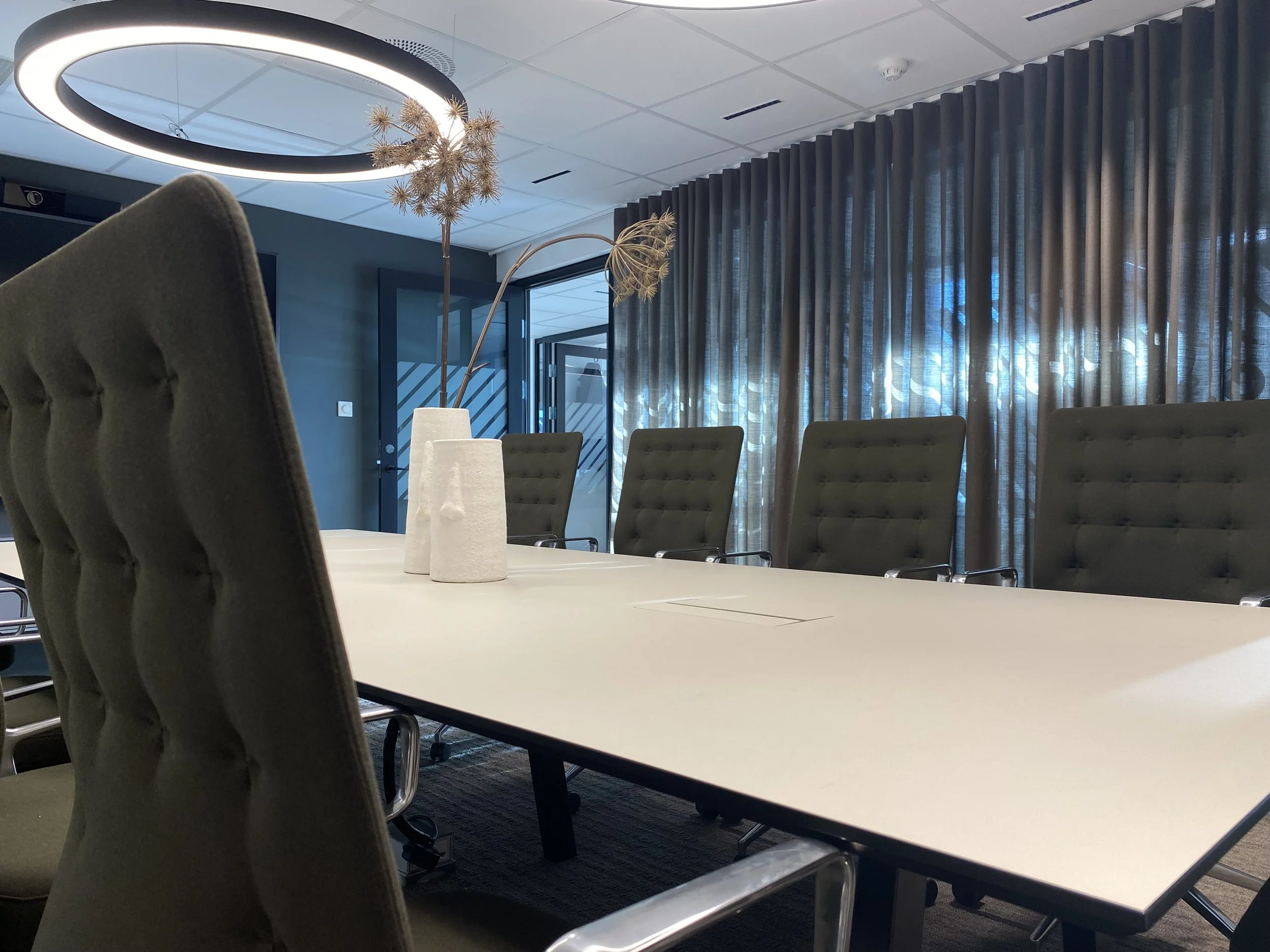 Hva er i dette bildet: et konferanserom med bord, stoler, lysarmatur og gardiner.