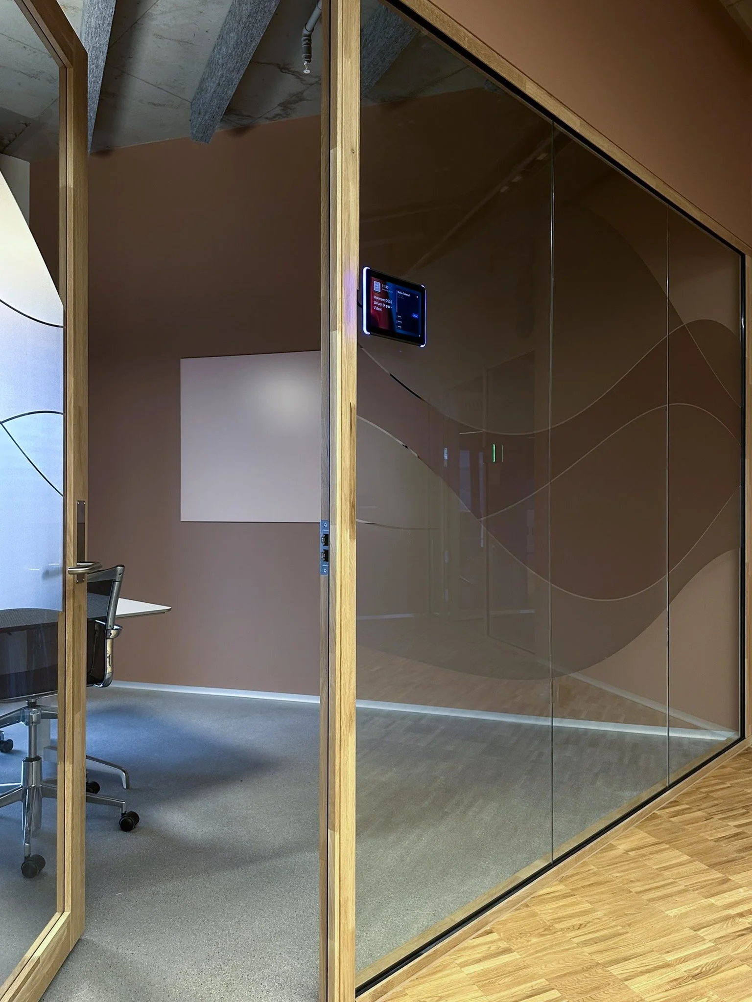 Et kontorlokale sett gjennom et glassvegg med treramme, som inneholder et lite møte- eller arbeidsrom med et bord, en kontorstol og en skjerm montert på veggen.